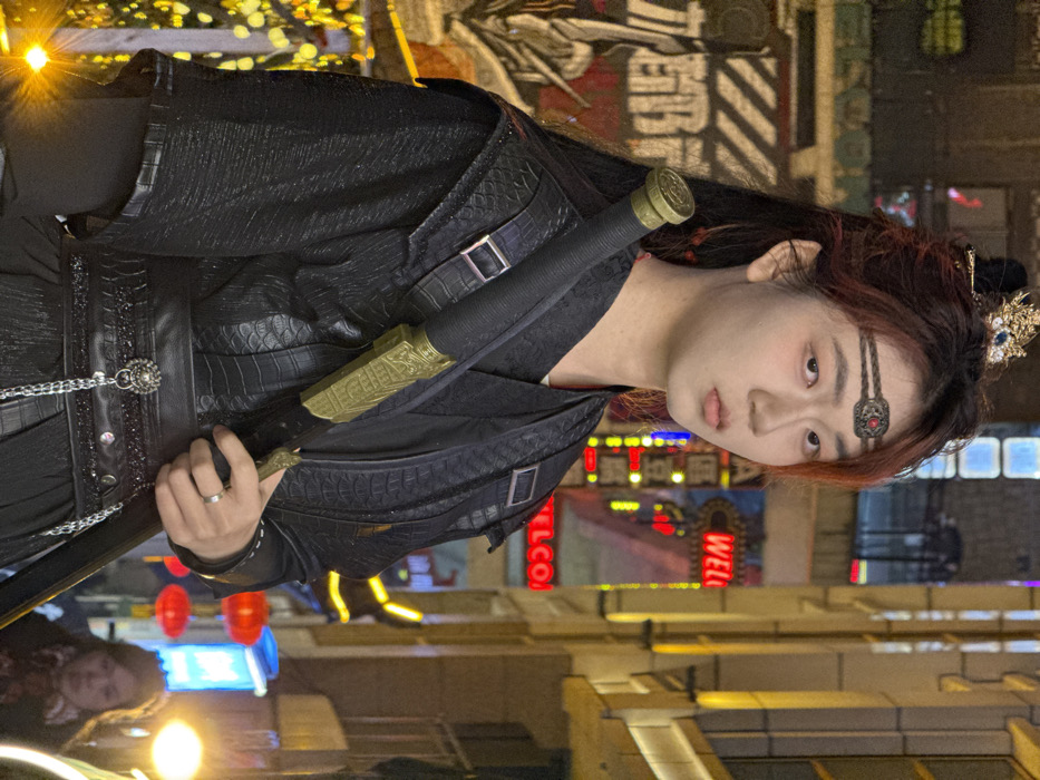 A young East Asian individual is centrally positioned in a medium shot, set against a blurred urban night environment in Yuzhong District, China. The individual has dark hair with reddish-brown highlights, styled with a gold-colored crown-like headpiece and a braided headband adorned with a central red gem. Their facial expression is neutral, with a direct gaze toward the viewer.
They are attired in a dark, multi-layered costume. The outer garment features a textured pattern, possibly mimicking reptilian scales, with shoulder straps and silver buckles. A wide black belt with a silver-colored animal-head buckle and attached chains is worn around the waist. A silver band-style ring is present on the ring finger of the left hand. The individual holds a sheathed sword diagonally across their body with their left hand, gripping the black-wrapped hilt and an ornate gold-colored section of the scabbard.
The background displays a brightly illuminated street scene at night. To the left, a large sign with visible Chinese characters and graphics is partially illuminated. To the right, blurred modern architectural structures with illuminated windows and vertical light elements are visible. Numerous neon lights, including a red sign partially displaying "WELCOME," are present. Red lanterns are faintly discernible in the lower right background, where a blurred figure of another individual is also visible. The scene is lit by various artificial light sources, creating a high-contrast nocturnal atmosphere.