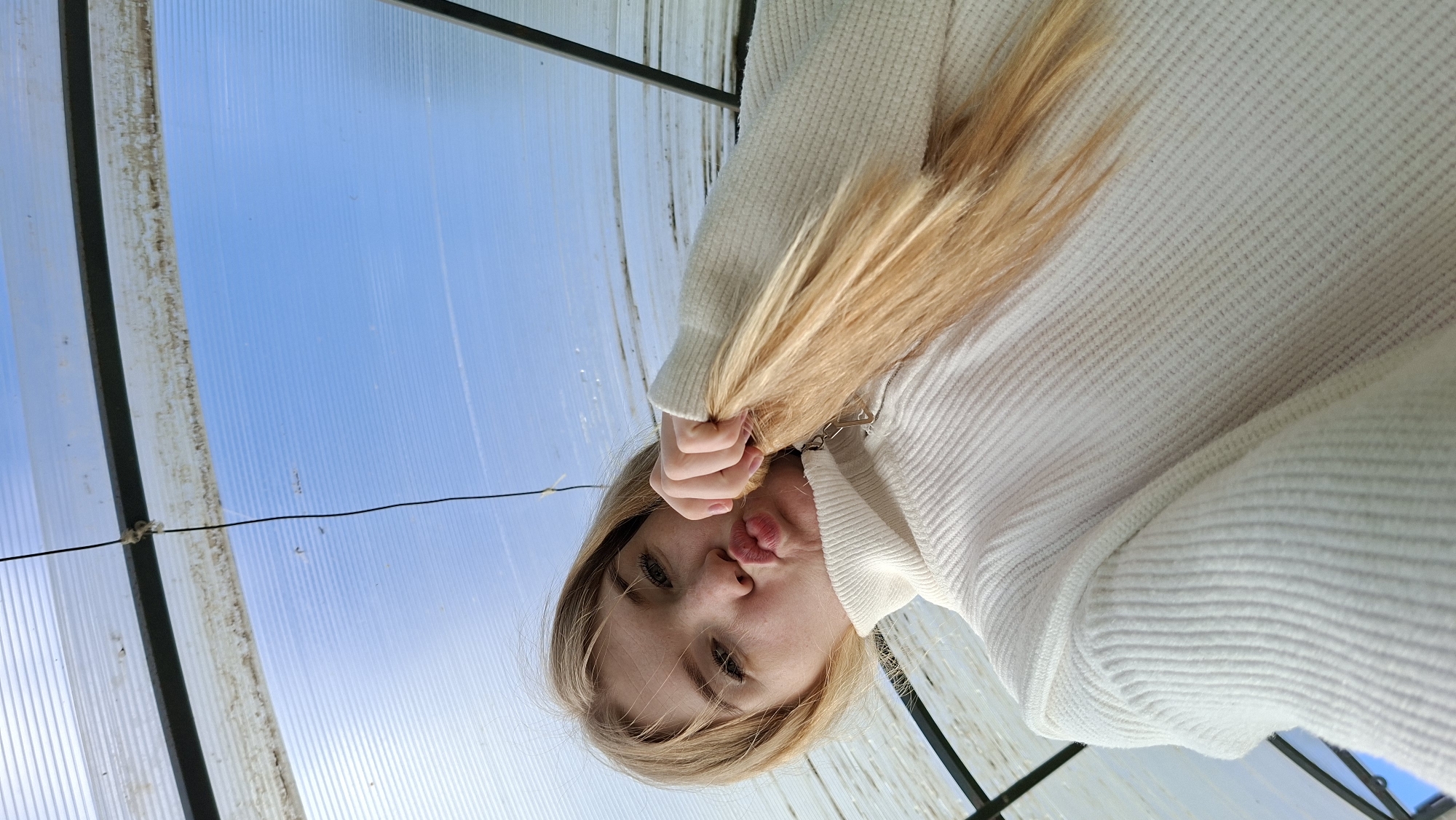 A low-angle, selfie-style portrait captures a young person with long blonde hair, looking upwards towards the camera. She is wearing a light-colored, ribbed knit sweater and makes a playful "duck face" with puckered lips. Her hand is visible, holding a section of her hair.
The background consists of translucent, corrugated panels, likely plastic or polycarbonate, through which a clear, bright blue sky is visible, indicating daytime with good weather. Dark structural beams or frames are also present, some exhibiting dirt or discoloration along their edges. A thin, dark cable or wire stretches horizontally across the scene. The setting appears to be inside or under a greenhouse, solarium, or similar covered structure, located in Большой Букор, Russia. The individual's expression conveys a lighthearted and playful mood. There is no visible text in the image.
