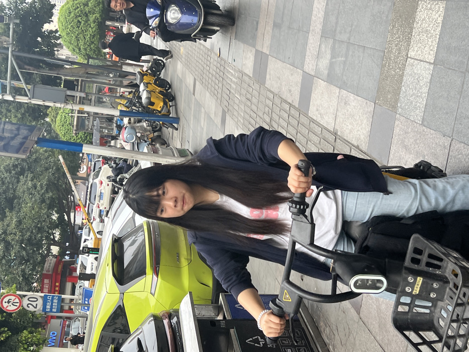 This image captures an urban street scene in Jiangjin, China, under overcast conditions.
In the immediate foreground, a female with long, dark hair and bangs is seated on a black electric scooter/bicycle on a grey tiled sidewalk. She wears a dark blue zippered hoodie, a white t-shirt with pink text, and light blue jeans, with her hands gripping the handlebars. Her mouth is slightly pursed. A black basket is affixed to the front of the scooter. To her left, a metallic utility box with a recycling symbol is partially visible.
In the midground, a lime-green SUV is parked behind the female, facing left. On the paved road, several other vehicles, predominantly white, are parked. A white traffic barrier arm extends over the road. A pole displays a red and white circular "no pedestrian" sign marked "20", and a blue sign with Chinese characters that read "请出入口" (meaning "Please enter and exit"). Further right on the sidewalk, multiple yellow shared electric scooters are parked. A man in a dark jacket walks away from the viewer on the sidewalk. On the paved road, another man, also in a dark jacket, drives a dark blue scooter/motorcycle towards the right.
The background includes commercial buildings with "VIVO" and "彩票" (lottery) signage, and green foliage from trees.