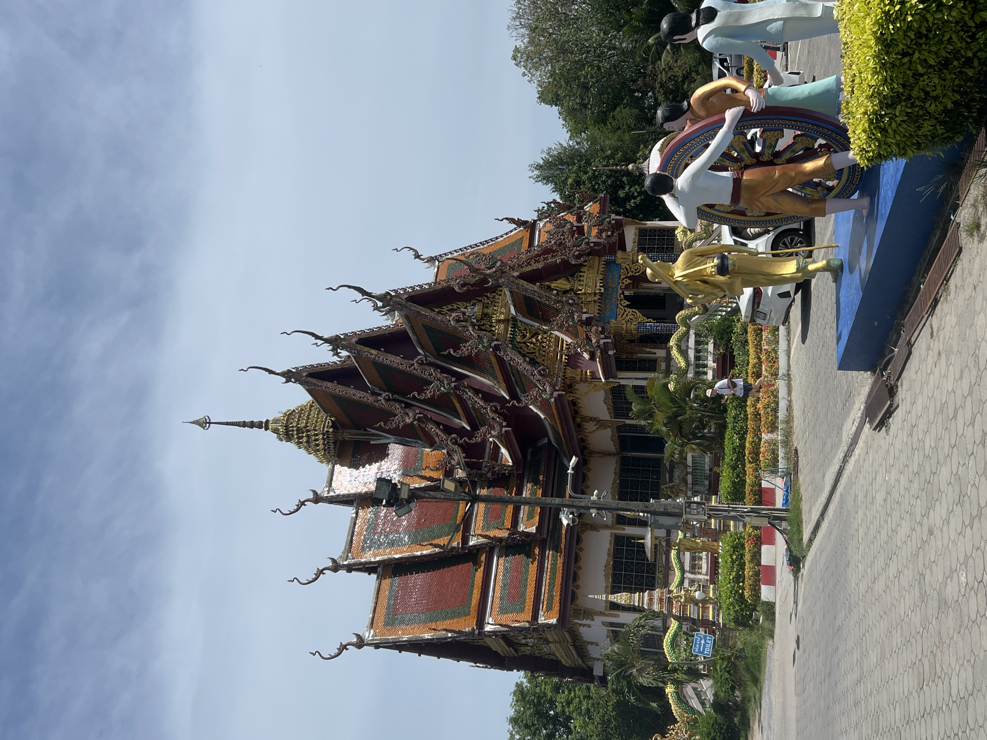 The image shows a temple in Baan Plai Laem, Thailand. The temple has intricate rooftops with gold embellishments and colorful tiles. Next to the temple are statues of people operating a wheel, and a golden statue of a figure with a weapon. A paved walkway is adjacent to the statues, with trees and greenery in the background. A person is visible in the background between the temple and the statues. Cars are parked nearby. The sky is partly cloudy.