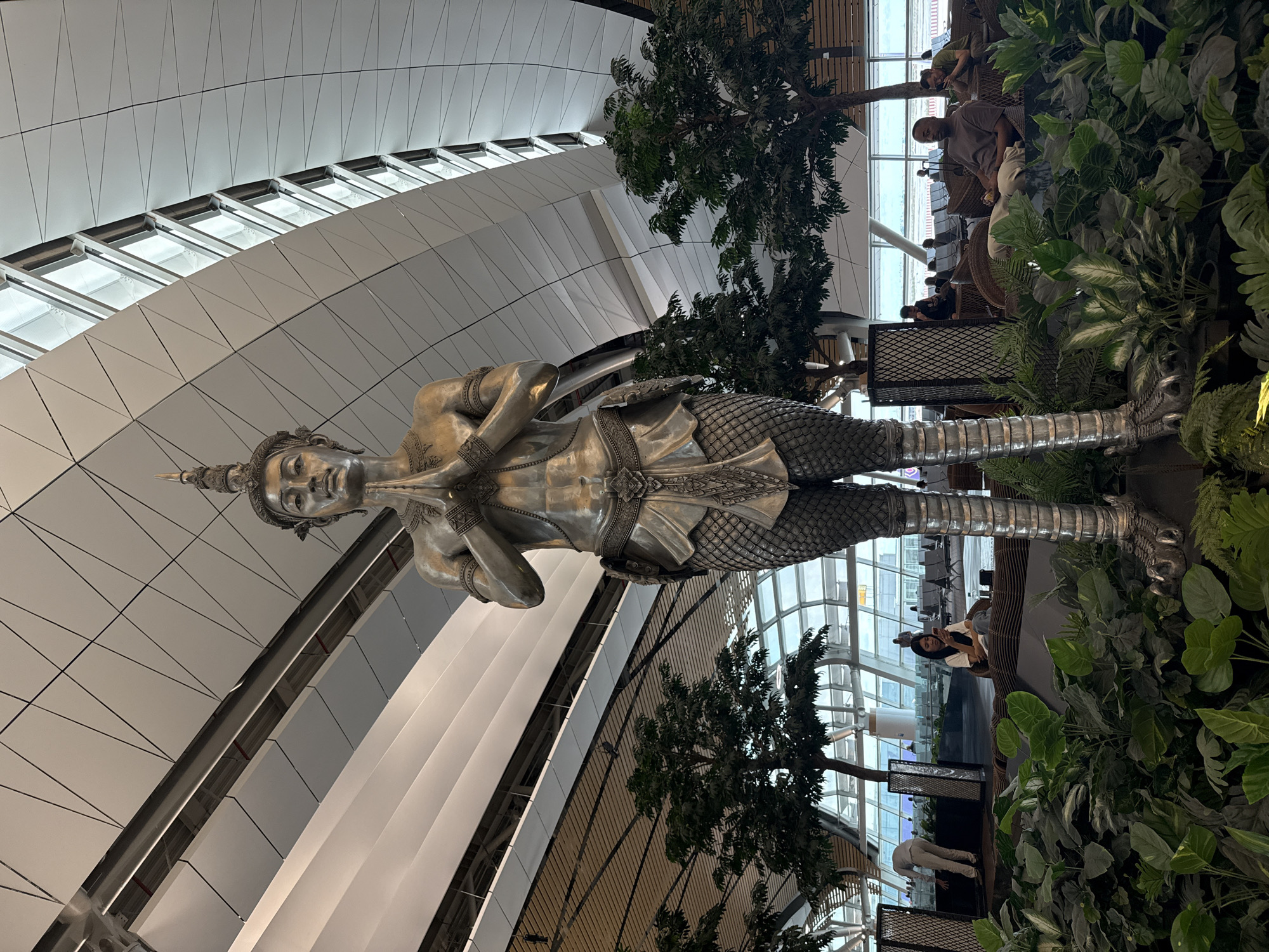 The image depicts a large, silver-colored statue of a Kinnaree, a mythical creature in Thai folklore, at the Suvarnabhumi Airport in Nong Prue Subdistrict, Bang Phli District, Samut Prakan Province, Thailand. The statue has a human-like upper body and a bird-like lower body, with intricate detailing in the scales, jewelry, and facial features. It stands amidst lush greenery with trees and various plants. In the background, passengers are seen relaxing on seats near windows overlooking the airport grounds. The ceiling features a modern design with geometric patterns and windows providing natural light.