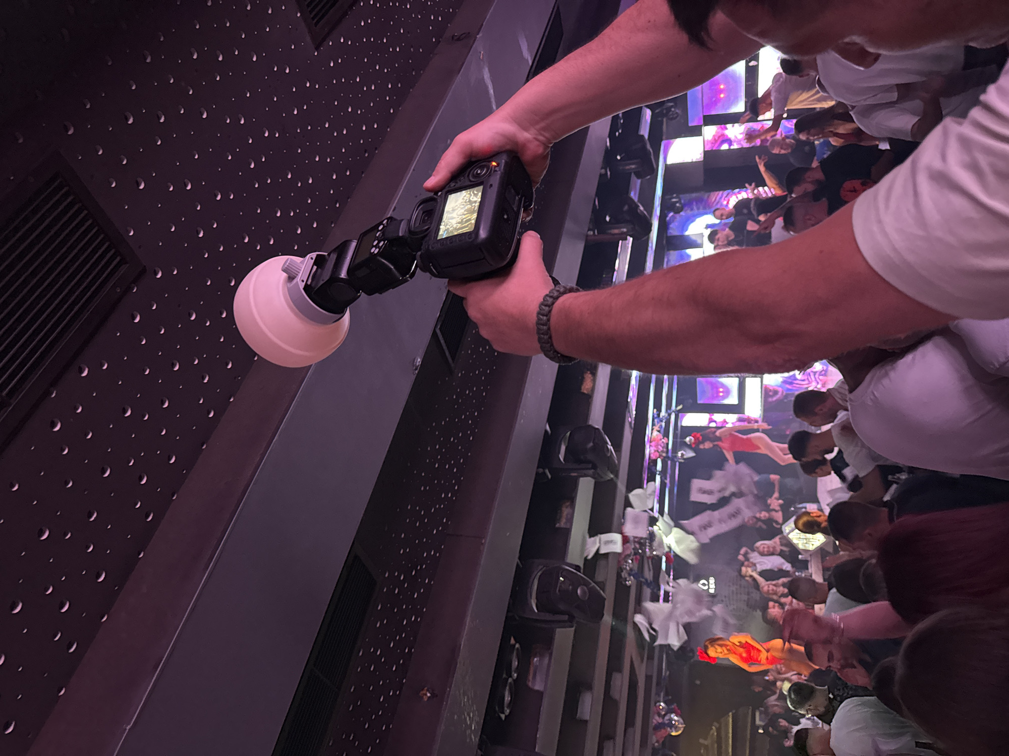 A photographer holds a camera with a flash attachment, taking a photo of a crowd of people in a club. The crowd appears to be celebrating, possibly at a party or event. Decorations and displays are visible in the background. The scene is indoors. I am unable to confirm the location in Blagoevgrad, Bulgaria.