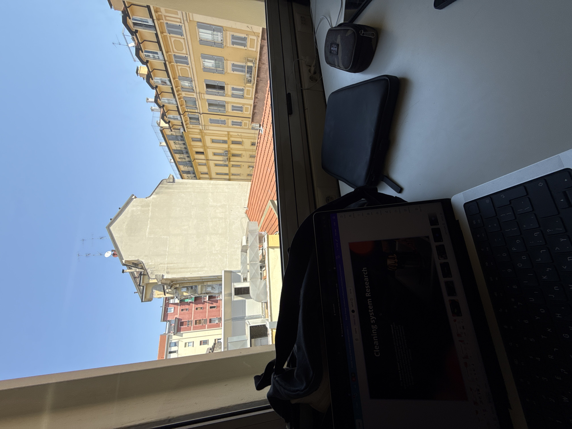 This image depicts a view from a window in Milan, Italy. Visible outside is a clear blue sky with several buildings, including a tall yellow building with rows of windows, a white building, and red buildings in the distance.
Inside, on what appears to be a desk or window ledge, there's a laptop displaying a presentation slide titled "Cleaning system Research." A black camera case, a black laptop case, and part of a keyboard are also on the desk surface. The scene suggests someone working from home with a view of the city.