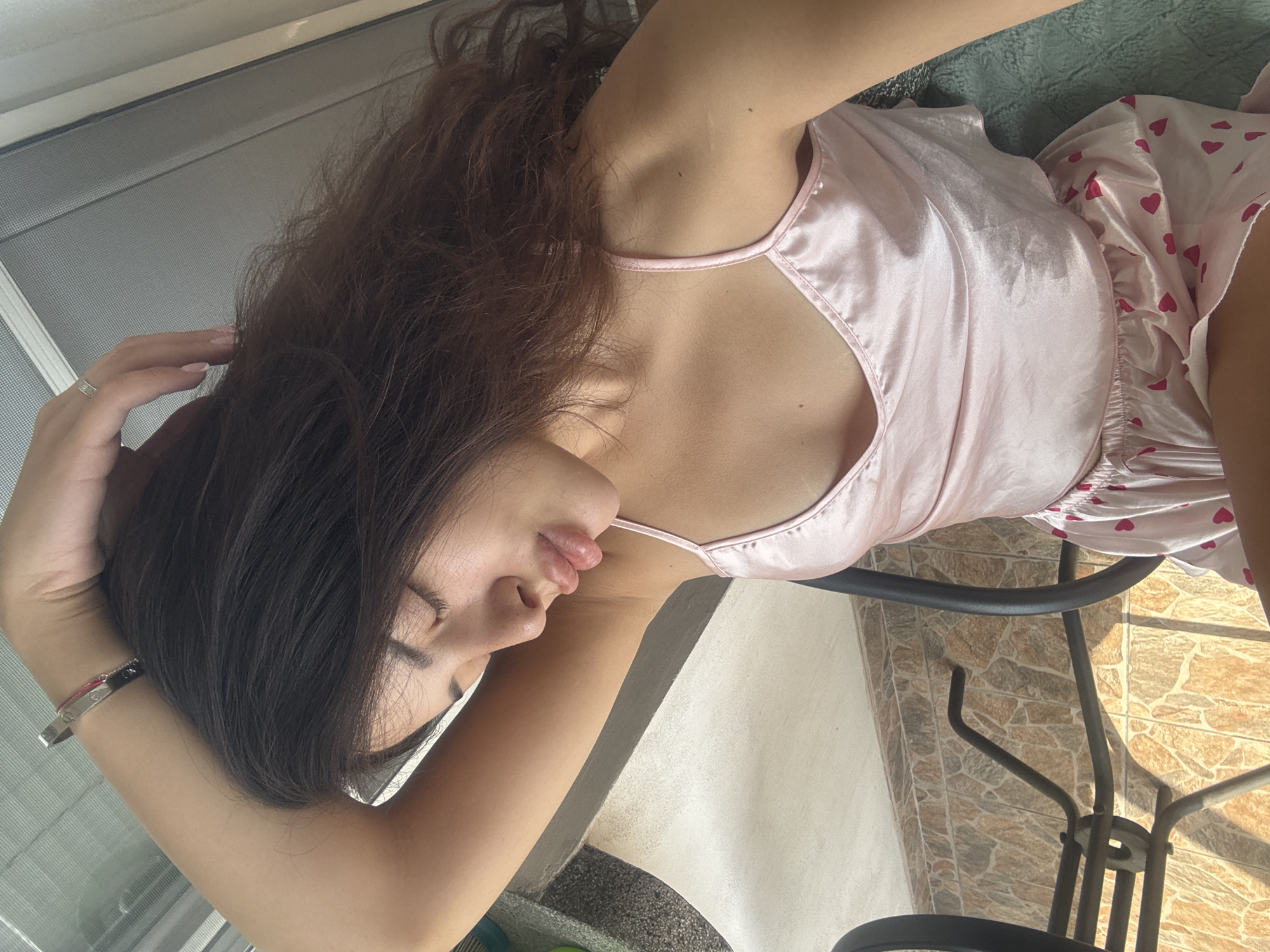 A young woman with dark brown hair lies on a chair, her head resting on her arm. She is wearing a light pink satin camisole with delicate straps and matching pink heart-patterned shorts. Her eyes are closed, and her lips are slightly parted. A silver bracelet adorns her left wrist, and a ring is visible on her right hand. The background suggests an outdoor setting, possibly a balcony or patio, with tiled flooring and a blurred glimpse of a table and chair. The lighting indicates daytime.