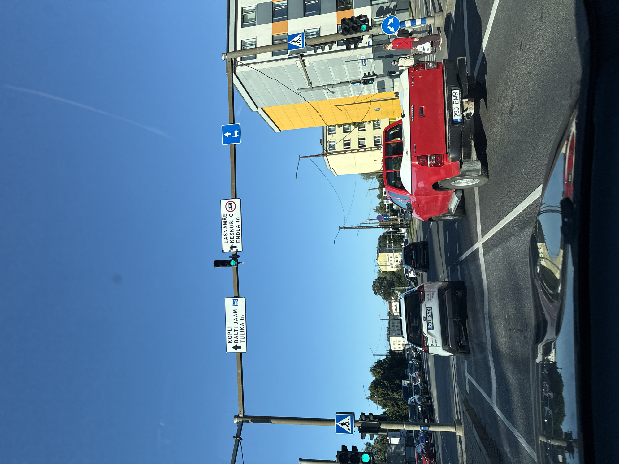 The image shows a street scene in Tallinn, Estonia, under a clear blue sky. In the foreground, a portion of a car's dashboard and windshield is visible, reflecting the surrounding environment. On the road, several vehicles are present, including a white SUV with license plate "731 SYX" and a red pickup truck with license plate "290 BMR". Traffic lights are visible, with the green light illuminated. Road signs indicate directions to "KOPLI BALTI JAAM TULIKA tn" and "LASNAMÄE KESKUS, C. ENDLA tn". A pedestrian crossing sign is also visible. In the background, buildings with yellow and gray facades line the street, and trees are present. Several pedestrians are seen on the sidewalk near a traffic light pole. The scene depicts a typical urban environment with traffic and infrastructure.