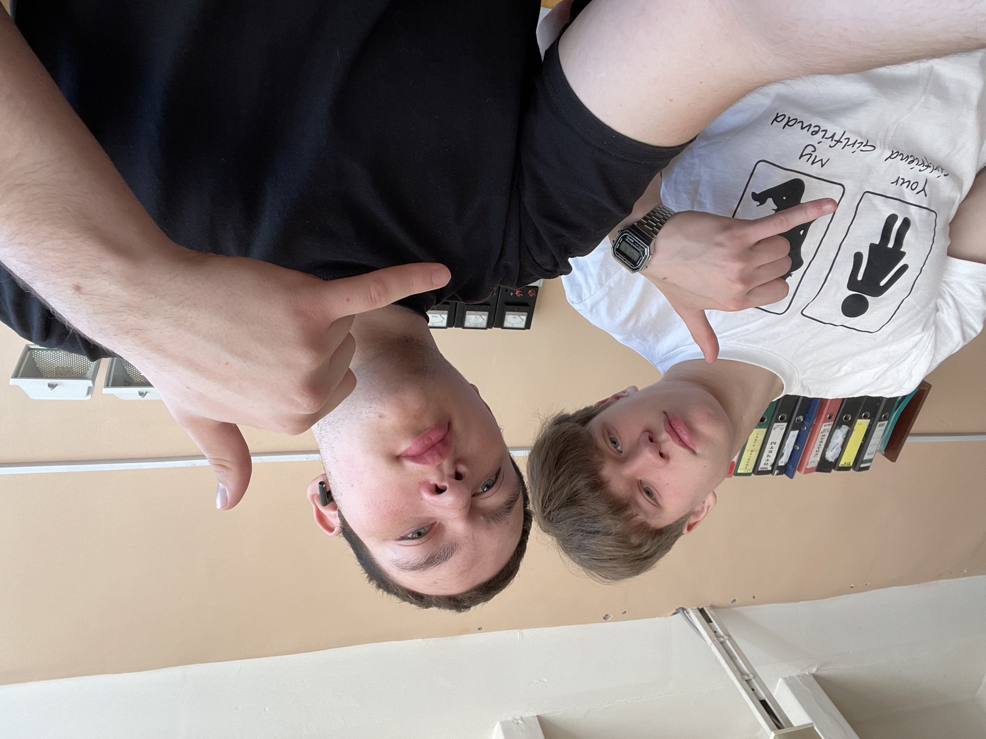 Two young men are positioned close to the camera lens, looking down at the viewer. The man on the left is wearing a black t-shirt and has a black wireless earbud in his left ear. The man on the right is wearing a white t-shirt with a cartoon drawing showing "My girlfriend" next to a drawing of a dog, followed by "Your girlfriend" next to a drawing of a woman. He is also wearing a digital watch on his left wrist. Both men are giving the "thumbs down" gesture.
The background is a beige wall with some items attached to it, including some electrical equipment with gauges and a shelf containing several binders of various colors. The bottom portion of the image shows a white ceiling with a lighting fixture and some sort of track or rail system.
The location can not be confirmed by the data provided.