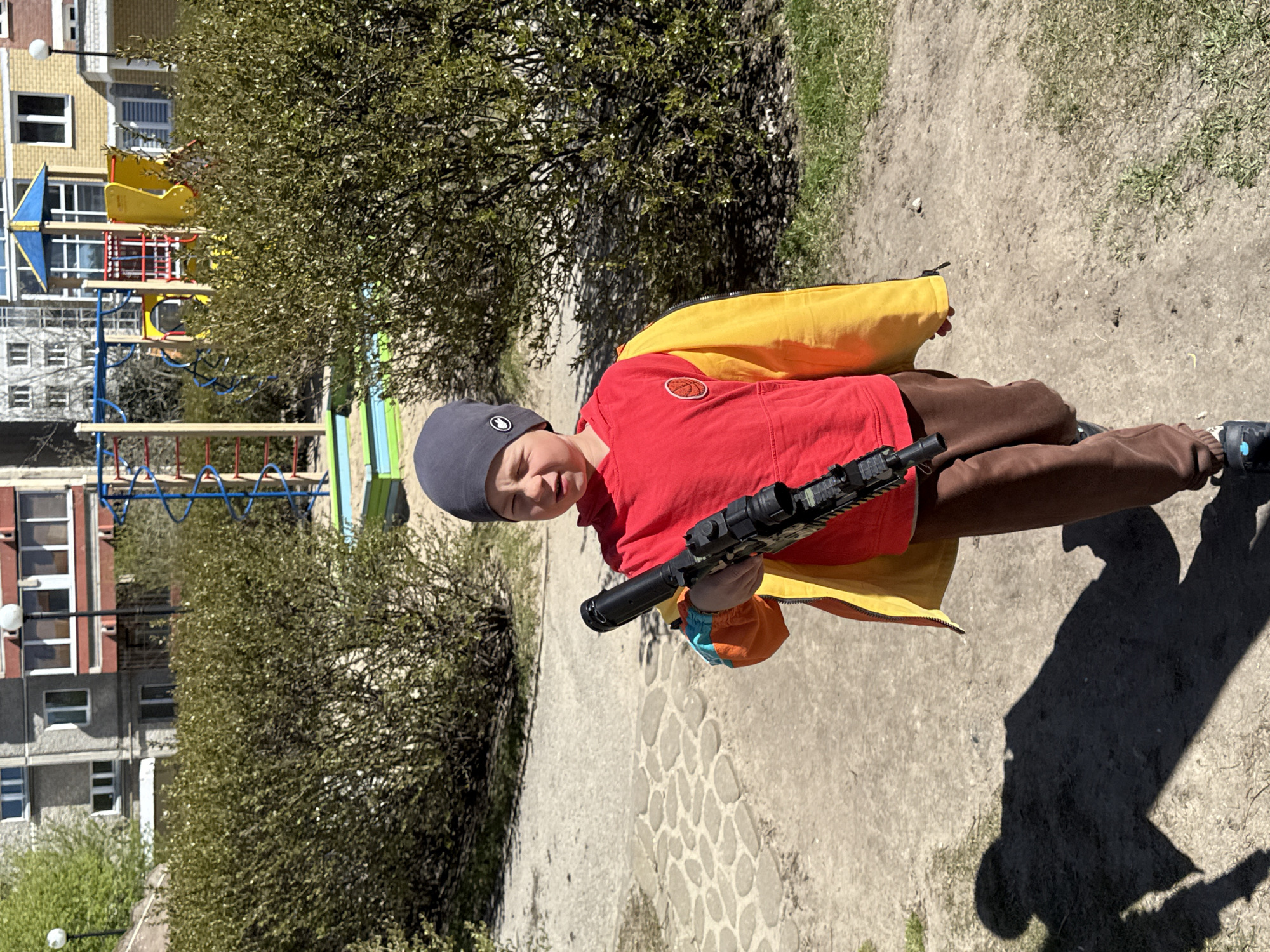A young boy in a gray knit hat, red t-shirt, yellow vest, and brown pants stands outdoors holding a black toy rifle. The setting appears to be a park or open space in an urban environment, likely Yekaterinburg, Russia, given the user's prompt. There are trees and bushes around, and a playground structure with slides and climbing equipment can be seen in the background, as well as multiple-story residential buildings. The ground is a mix of dirt, concrete, and some grass. The boy's shadow is visible on the ground.