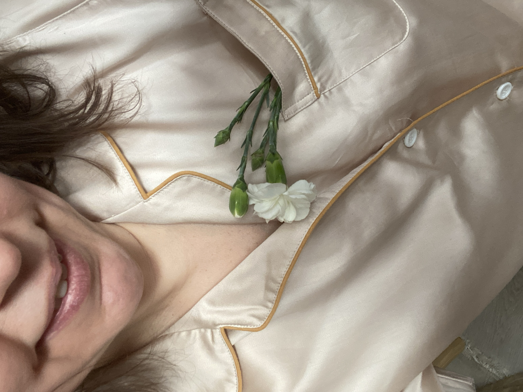 A close-up, overhead shot captures a person partially visible on the left, lying down on what appears to be a bed or soft surface. The lower part of their face, including the mouth, chin, and cheek, is visible, along with dark brown hair spilling onto a light-colored fabric. The person's mouth is upturned in a gentle, subtle smile.
They are wearing a light champagne or pale pink satin-like pajama top, which fills most of the frame. The pajamas feature a contrasting golden-brown piping along the seams, collar, and pocket edges. Two white buttons are visible on the right side of the garment. Tucked into the pajama top's breast pocket are several delicate green stems with multiple small white flower buds, and one fully bloomed small white carnation, adding a touch of natural beauty to the soft fabric.
The overall scene conveys a feeling of serenity and intimacy, possibly capturing a quiet moment of relaxation. There are no visible location cues within the image itself, though the provided context is Chaikovsky, Russia. No text is visible in the media file.
