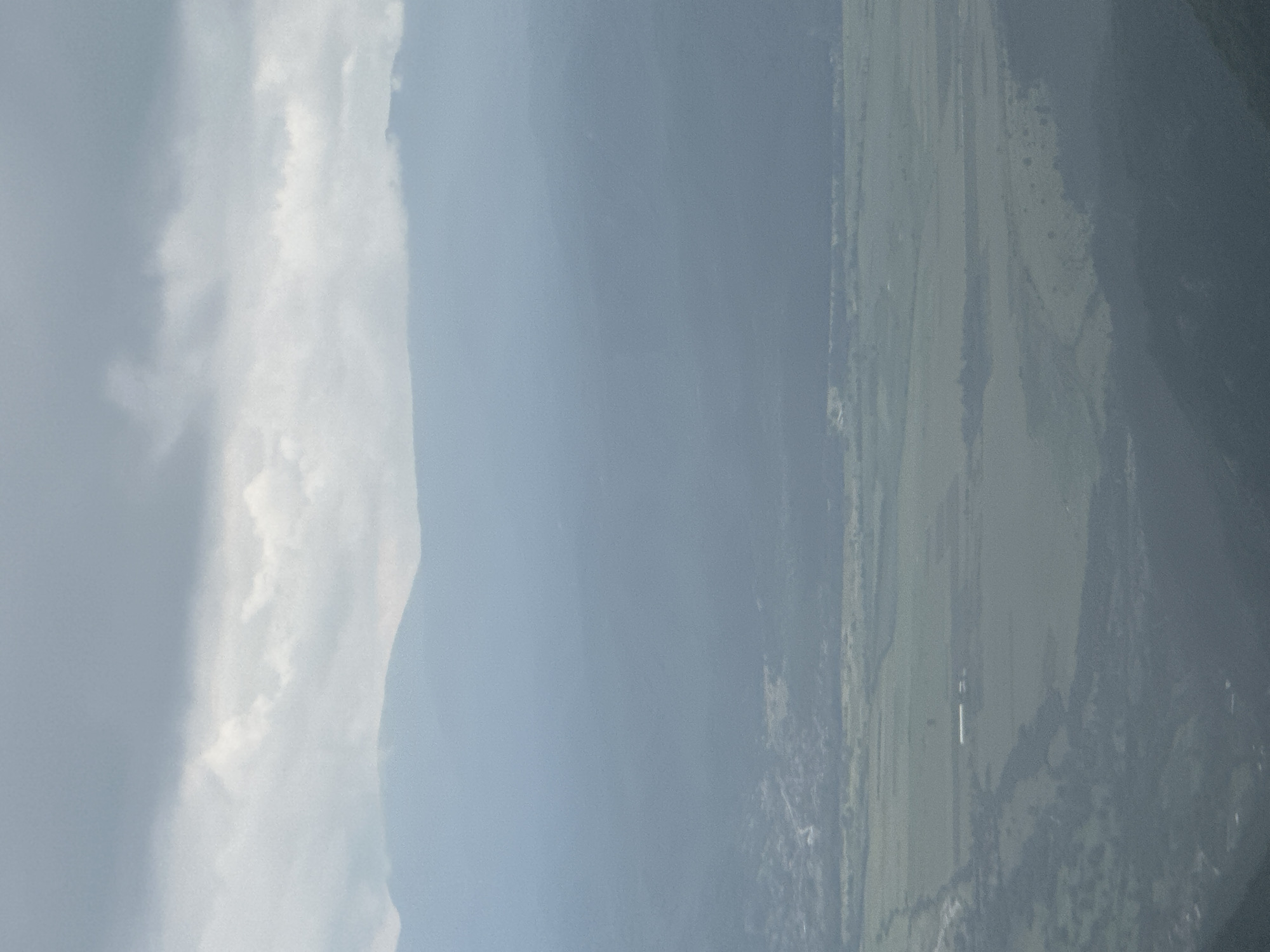 This is a high-angle or aerial photograph depicting a vast landscape, likely taken from an elevated position such as a mountain or an aircraft, in the region of Hristo Danovo, Bulgaria.
When viewed correctly oriented, the left and upper sections of the image are dominated by a partly cloudy sky, featuring prominent white and light gray cloud formations that stretch across the frame. Below the clouds, in the middle distance, a long, hazy mountain range extends horizontally, its slopes transitioning from lighter tones near the sky to darker, possibly forested, areas.
The lower and right portions of the image reveal expansive green plains, indicative of agricultural fields or pastures, interspersed with darker patches of vegetation or scattered trees. A small, distant white building or structure is faintly discernible within one of these fields in the lower right area.
The scene conveys a sense of peaceful expanse during daytime, with diffused natural light suggesting either an overcast sky or significant atmospheric haze. There are no people visible, no specific events taking place, and no discernible text. The image provides a panoramic view of Bulgaria's diverse topography, encompassing both mountains and agricultural lowlands.