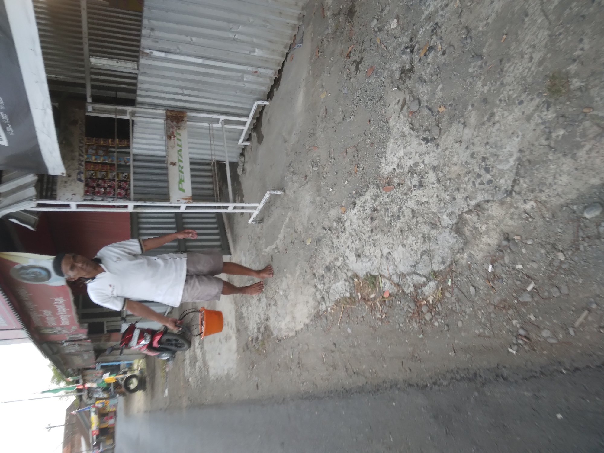 A middle-aged man with a dark complexion and a black peci hat is walking along a dirt and concrete path on the side of a paved road. He is wearing a white polo shirt with black trim on the collar and sleeves, and light brown shorts. In his right hand, he carries an orange bucket. His left arm is bent at the elbow and his left hand is by his side. He is barefoot.
To his left, a red and black motorcycle is parked on the side of the road. Behind the motorcycle, there are several small shops with red roofs and signs. One sign has text in red and white and appears to advertise "Durung Kapal Api".
To the man's right, there is a metal stand with shelves displaying various items, some of which appear to be packaged goods, possibly food or drinks. The stand has a sign with "PERTAMINA" and "PERTALITE" printed on it. Behind the stand, there is a corrugated metal wall.
The environment appears to be a roadside in a village or small town, possibly in Indonesia given the presence of the peci hat and the shop signs. The road is paved, and the path next to it is a mix of dirt, concrete, and gravel, with some dry leaves scattered on the ground. The lighting suggests it is daytime, possibly late afternoon or early morning, with overcast conditions.