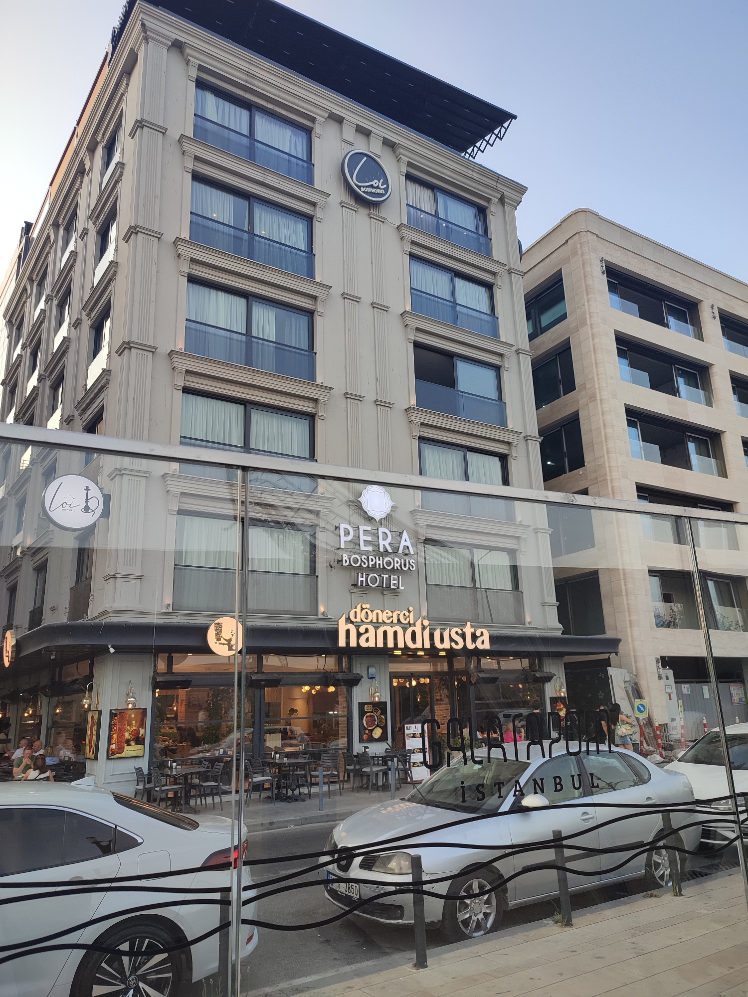 In Beyoğlu, Turkey, a multi-story building with ornate architectural details is prominently featured. The building has a light-colored facade with multiple floors, each featuring rows of windows with dark frames and balconies. A sign on the building reads "PERA BOSPHORUS HOTEL." Below the hotel, at street level, is a restaurant with a glass storefront. Inside, tables and chairs are visible, and people are seated and moving about. Several cars are parked along the street in front of the restaurant and hotel. The scene appears to be captured during daylight, with clear skies overhead.