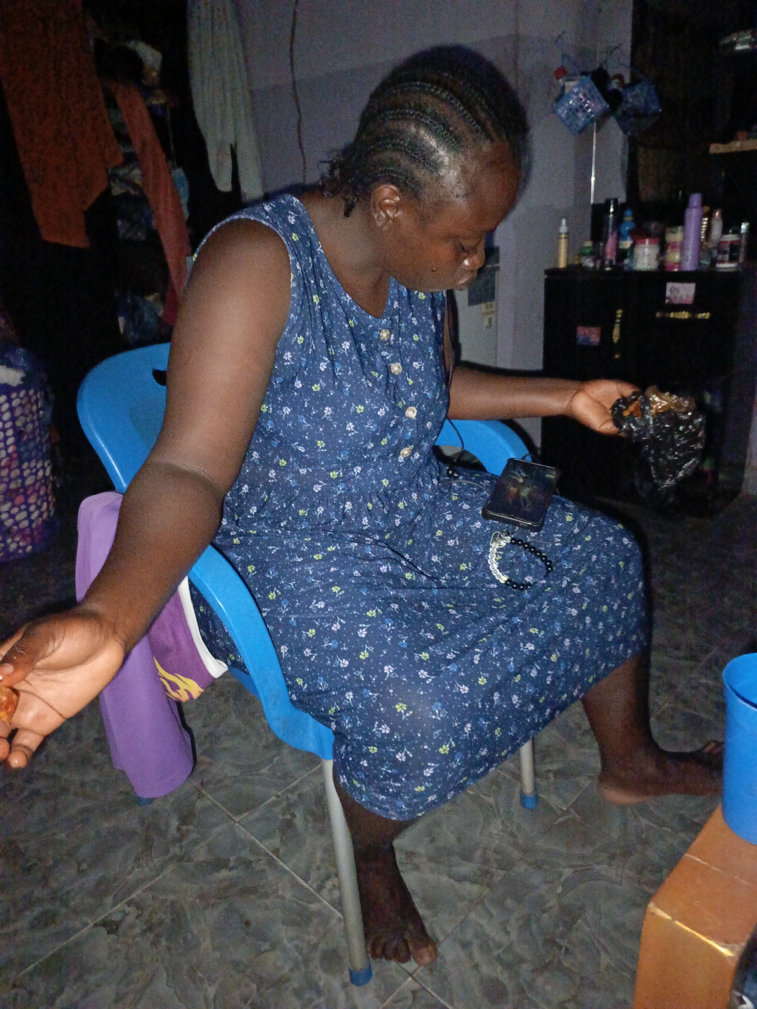 A woman with braided hair, wearing a floral print dress, sits on a blue plastic chair indoors. In her left hand, she holds a shiny, dark object, and a cell phone rests on her lap along with a beaded bracelet. The room features tiled flooring, a dark cabinet with various items on top, and clothing hanging in the background. A purple item is draped over the back of the chair, and a blue bucket sits on the floor near the chair. The city is Unknown, Unknown.
