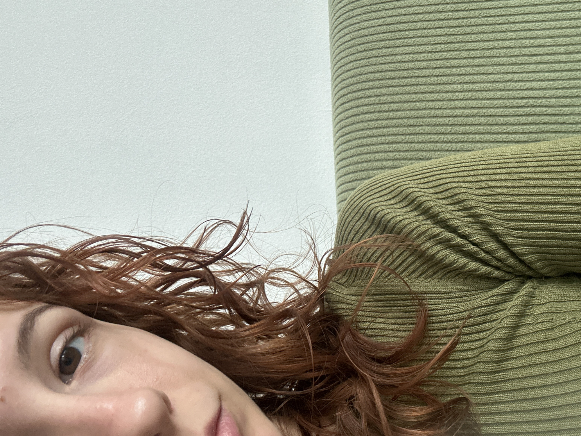 A close-up, eye-level shot depicts a person lying down. Only part of their face is visible, including their eye, nose, and part of their mouth. They have auburn, wavy hair. The person wears a ribbed, light green shirt or sweater. Behind them is a white wall. The location is unknown. The image does not allow for the determination of Novi Sad, Serbia, or any specific city.