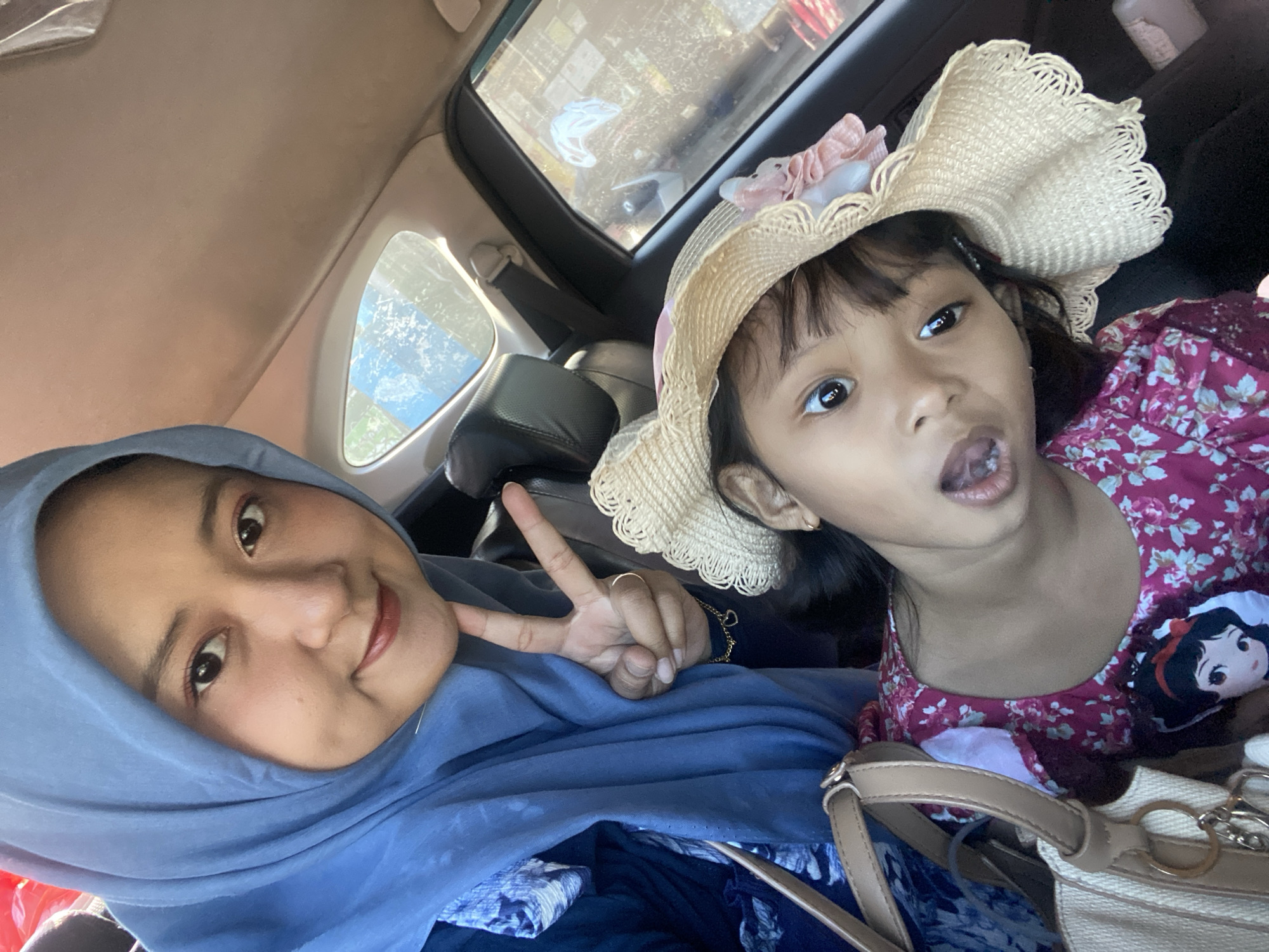 The image captures two females inside a car. The older female is wearing a blue hijab and making a peace sign, smiling at the camera. The younger female wears a straw hat adorned with a pink ribbon. Her expression is playful, with her tongue slightly sticking out. Both are situated in the back seat of the car. A window is visible in the background and a bag is in the lower right. The exact location is inside a car in Muktisari, Indonesia.