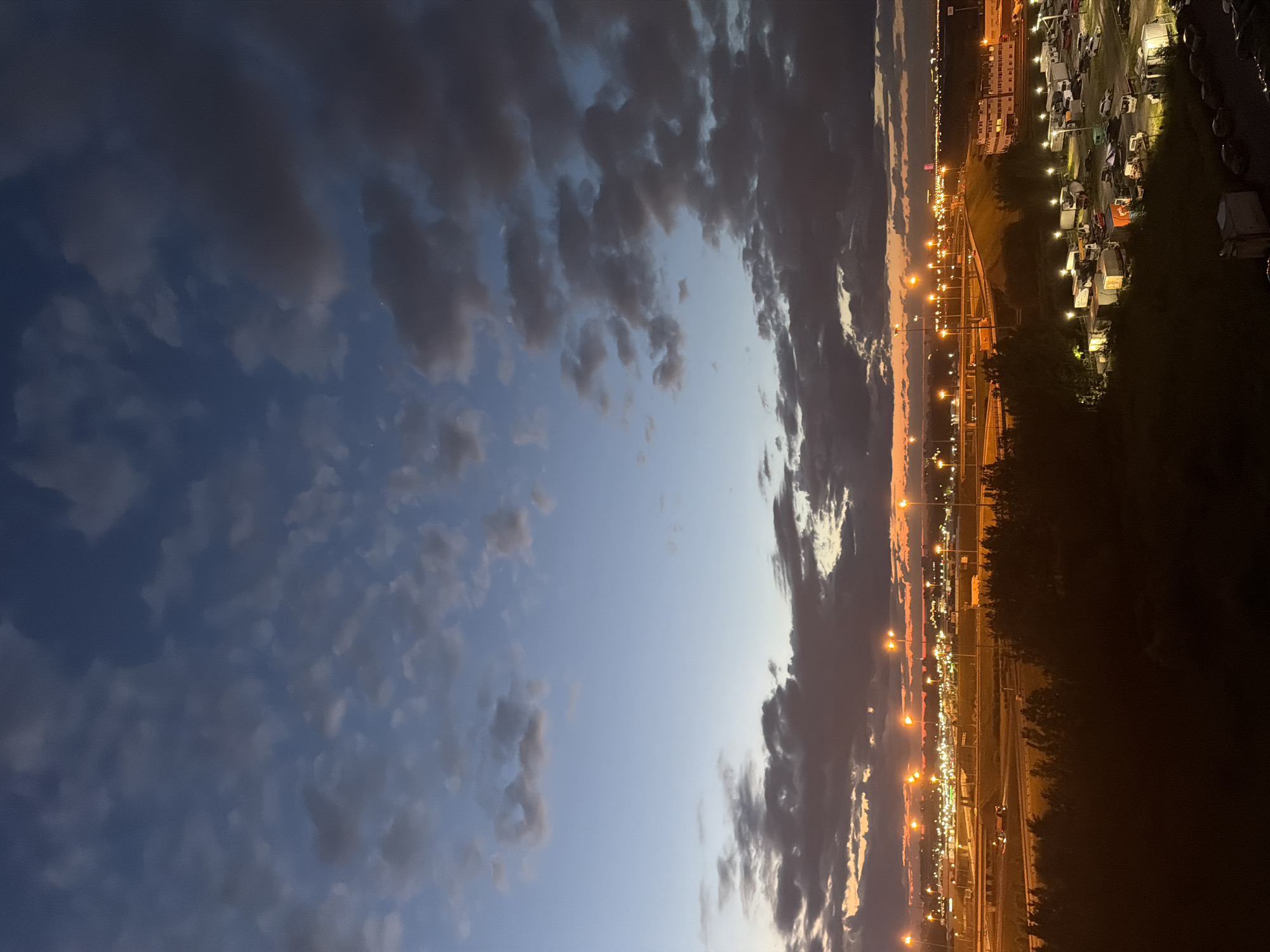 The image shows a twilight view of an industrial area, possibly near an airport or highway interchange, in the city of Unknown, Unknown. The sky is a blend of deep blues and oranges, with scattered clouds. Numerous streetlights and building lights illuminate the scene, casting a warm glow on the landscape. Several vehicles, including trucks and cars, are parked in a lot. Trees and dark foliage are visible in the foreground, obscuring the lower portion of the image. The overall impression is one of a bustling urban periphery at dusk.