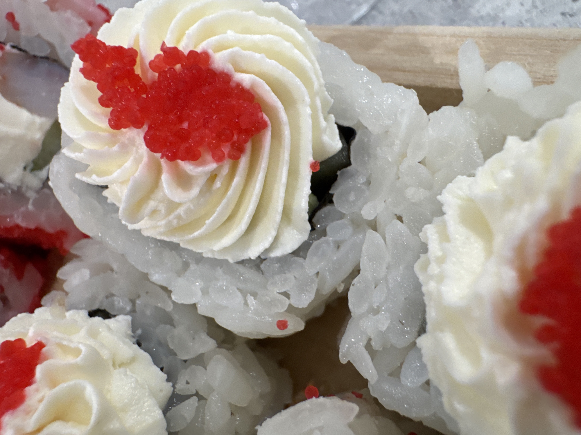 A close-up shot of several sushi rolls arranged in a wooden tray. The sushi rolls are primarily white rice with a swirl of white cream cheese on top. The cream cheese is decorated with red roe. The scene is a bright, even lighting, with no visible people. The sushi is the main subject, taking up the whole frame. The red roe is a small but noticeable detail. The environment is minimal, showing only the wooden tray. The location in the city of Noviy, Russia, is not evident from the image.