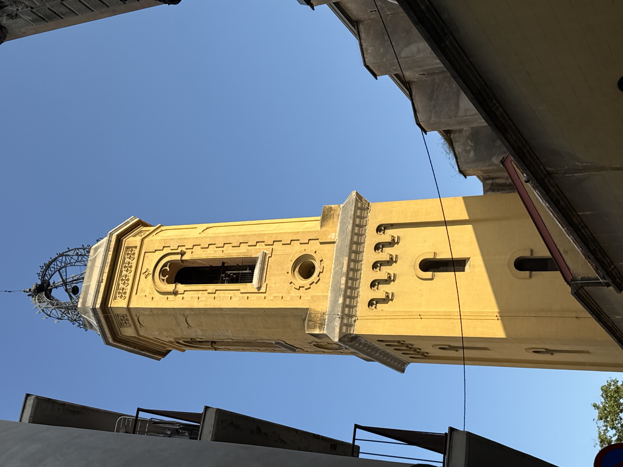 The image shows a low-angle shot of the clock tower, Torre del Rellotge, in Barcelona, Spain. The tower is made of yellow stone and is framed by other buildings on the sides of the shot. The tower is topped by an iron latticework structure. The sky is clear and blue.