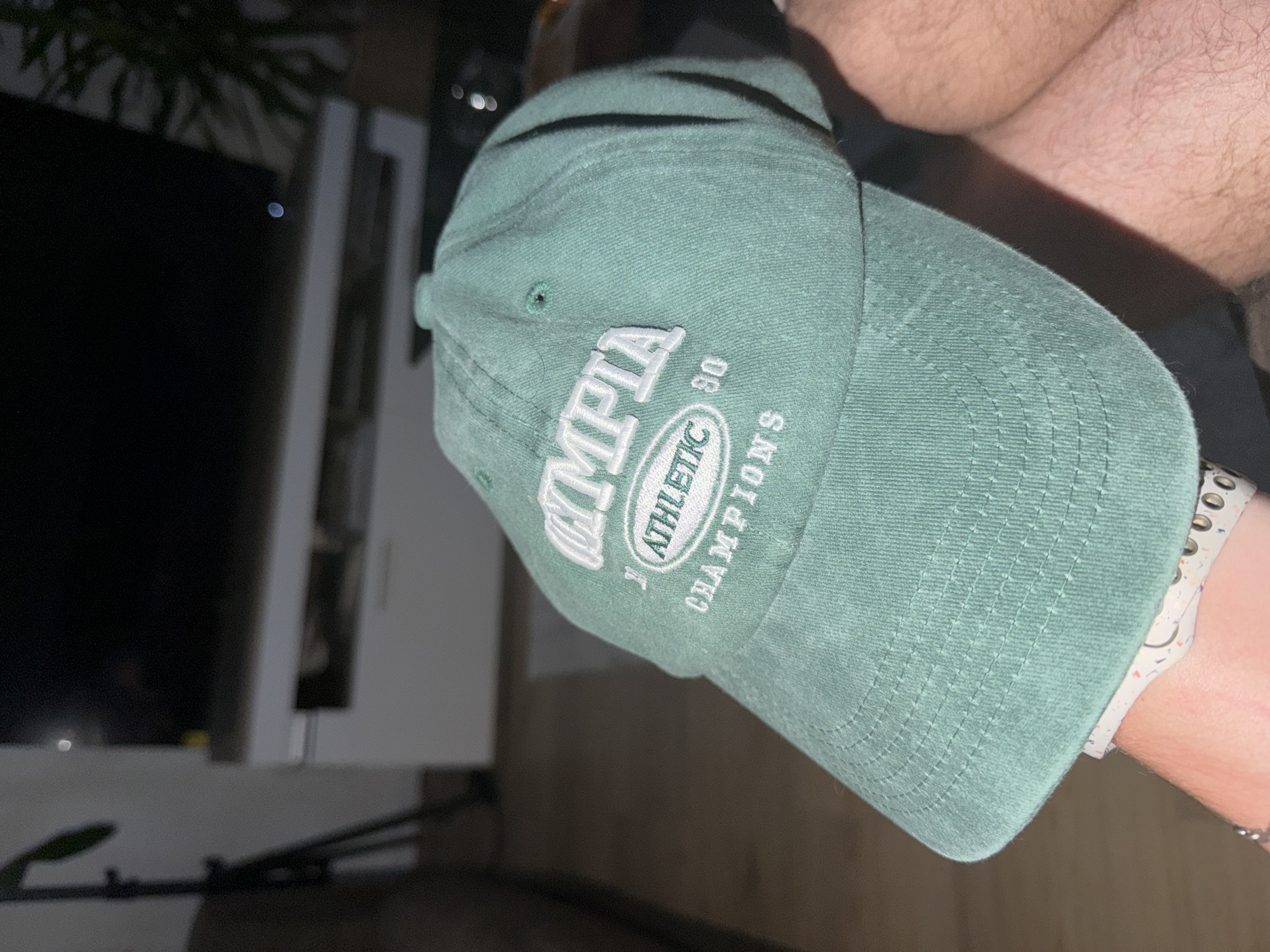 A close-up shot captures a green baseball cap with white embroidery. The embroidery on the front of the cap reads "OLYMPIA ATHLETIC '80 CHAMPIONS." The cap is held by a person's arm, part of which is visible in the lower right corner of the image. The background is out of focus, showing a dimly lit indoor setting with what appears to be furniture and possibly a television screen in the distance. The lighting suggests it is nighttime or a dark room.