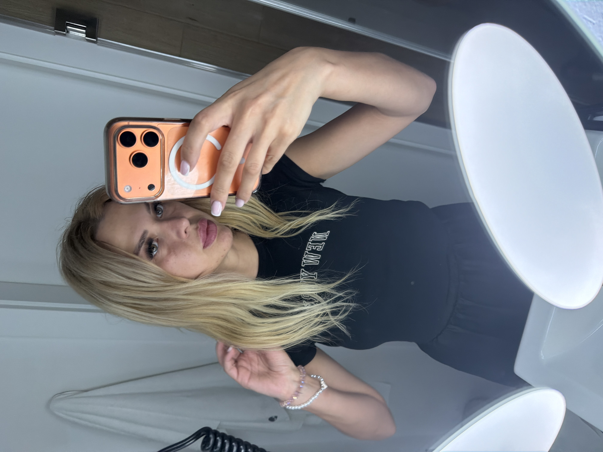A young woman with blonde hair and light makeup is taking a selfie in a mirror. She is wearing a black t-shirt with white lettering that appears to read "MEM YORK". On her wrist, she has a delicate bracelet with pink and white beads. The woman is holding up an orange iPhone in a clear case with a white circular magnetic attachment. Her nails are manicured with a light pink polish. She is looking directly at the camera, with her lips slightly parted.
The setting appears to be a bathroom or dressing area, indicated by the presence of a large, oval-shaped mirror with bright, illuminated edges. The background is mostly white and clean, suggesting an indoor environment. The lighting is bright, potentially from the mirror itself or overhead sources. The orientation of the photo suggests the woman might be in Polianytsia, Ukraine. There are no strong indicators of the time of day or weather conditions. The overall mood seems casual and self-aware, typical of a selfie. The reflection in the mirror shows the woman from the chest up, along with parts of the room and the phone she is using.