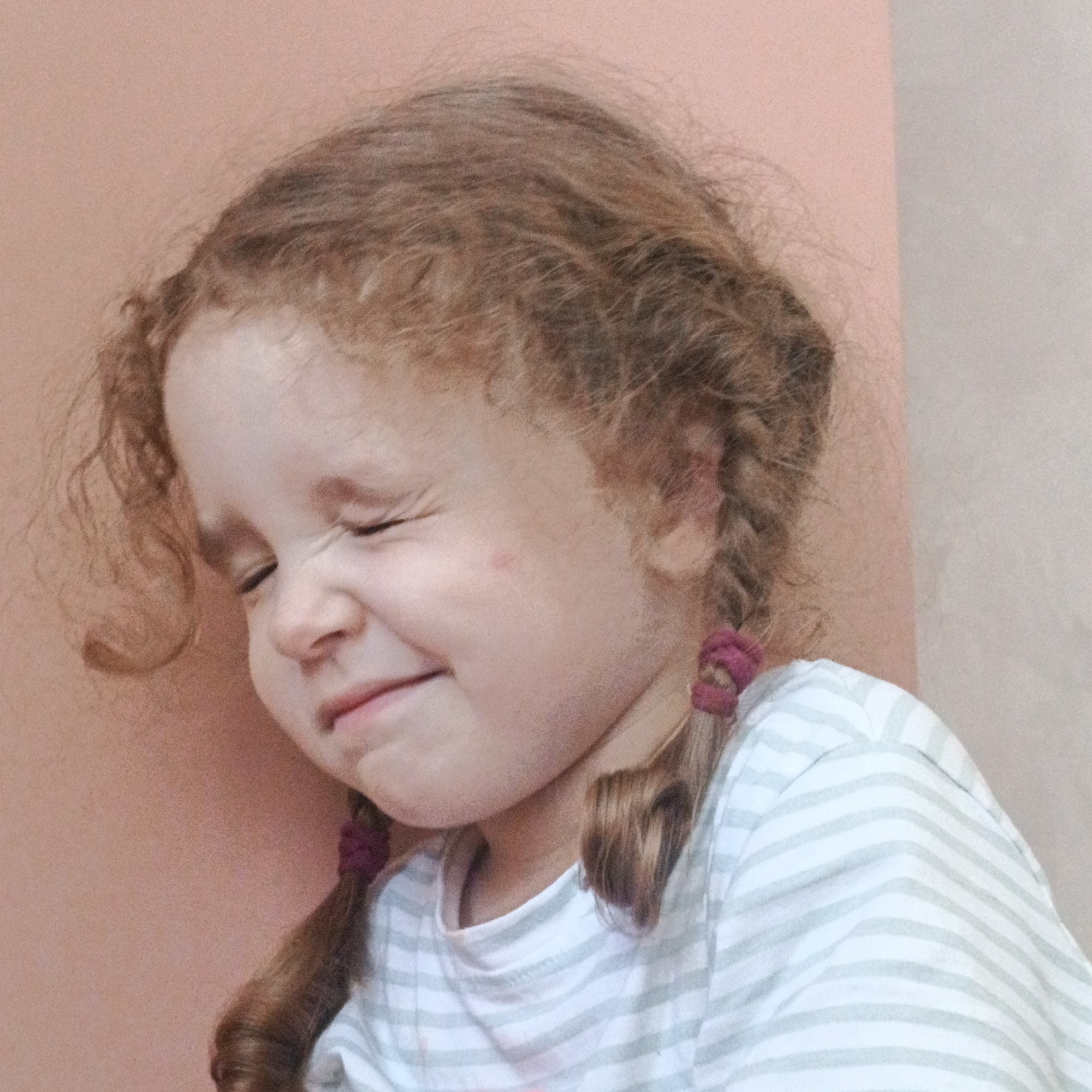 The image is a close-up, medium-shot photograph of a young child with reddish-brown, curly hair pulled into two pigtails secured with pink elastics. The child is wearing a light-colored t-shirt with thin, horizontal grey stripes. The child's eyes are squeezed shut, and their mouth is slightly open in an expression that suggests they are either laughing, grimacing, or experiencing discomfort. A small red mark is visible on the child's left cheek. The background is a soft-focus, two-toned wall, with the left side a peachy-pink color and the right side a lighter, neutral tone, suggesting an indoor setting. The image does not contain any information that could indicate its location in Марковский, Russia.