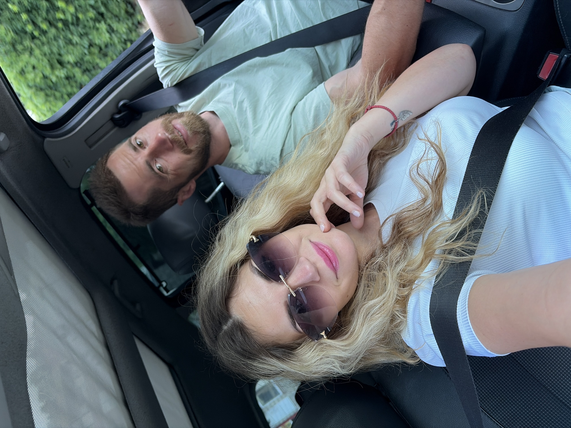 A man and a woman are in a car, possibly on a road trip. The woman is in the foreground, taking a selfie, and she's wearing large, fashionable sunglasses and a white top. Her blonde hair is styled in loose waves, and she has on bright pink lipstick. A red beaded bracelet with a heart-shaped charm is visible on her wrist. The man is in the background, driving. He has short brown hair and a beard, and he's wearing a light green t-shirt. Both individuals are wearing seatbelts. The car's interior is visible, including the dashboard and a sunroof. Outside the car, greenery suggests they are in a rural or suburban area, possibly near Budva, Montenegro, given the presence of houses and lush vegetation.