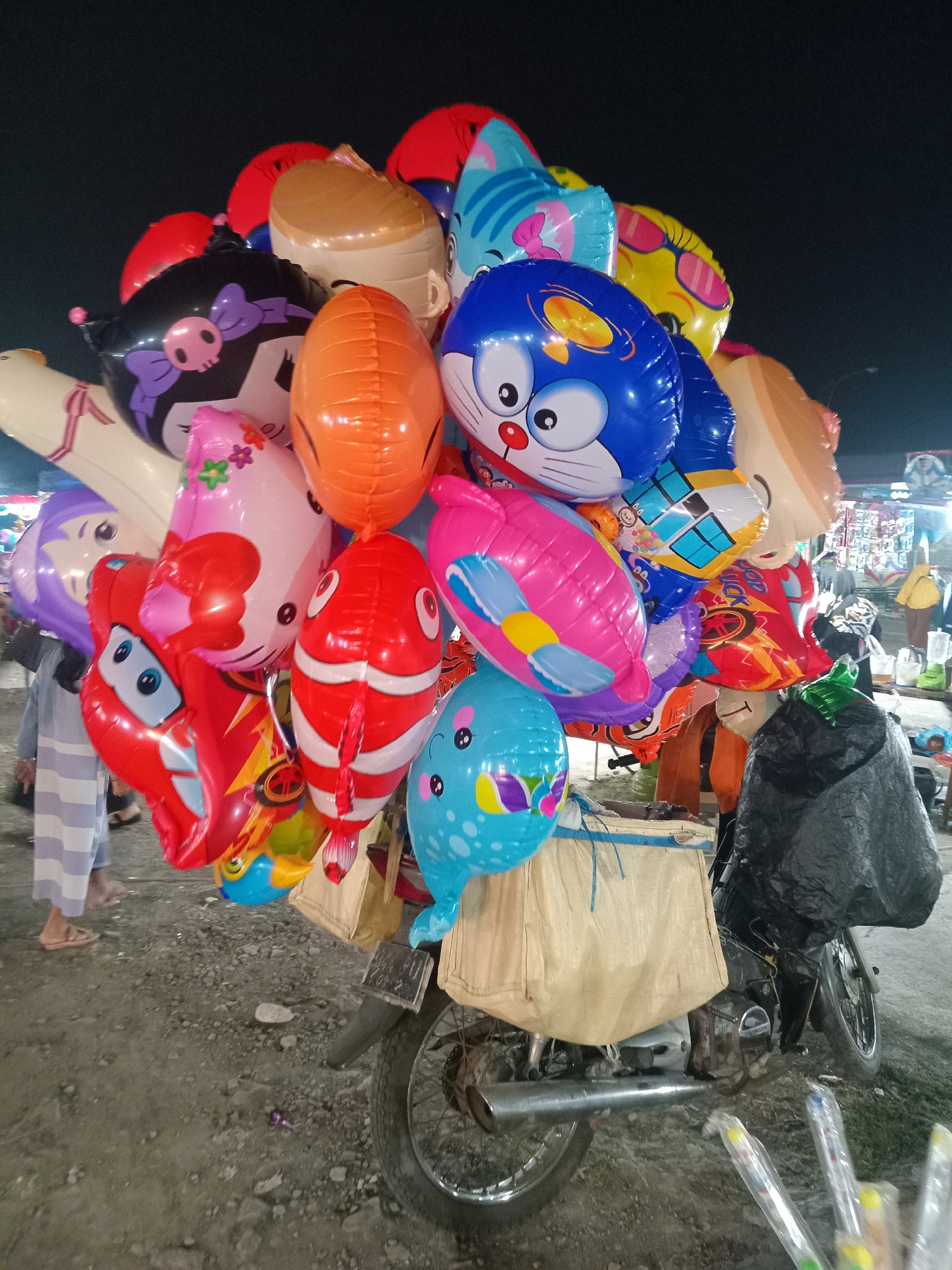 A nighttime outdoor market scene in Sedati Agung, Indonesia, features a motorcycle laden with a large bunch of colorful, character-shaped inflatable balloons. The balloons are predominantly cartoon-themed, including characters resembling Doraemon, Hello Kitty, and a clownfish. They are tied together and extend upwards and outwards from the motorcycle's rear cargo area, which is covered by a large, beige fabric bag. A black plastic bag is also draped over the rear of the motorcycle. The ground appears to be a mix of gravel and dirt. In the background, market stalls are visible, with some lights illuminating the area. Several people are present in the market, including a person in a striped robe and sandals walking in the left foreground and others in the background browsing stalls. The overall atmosphere suggests a lively local market event.