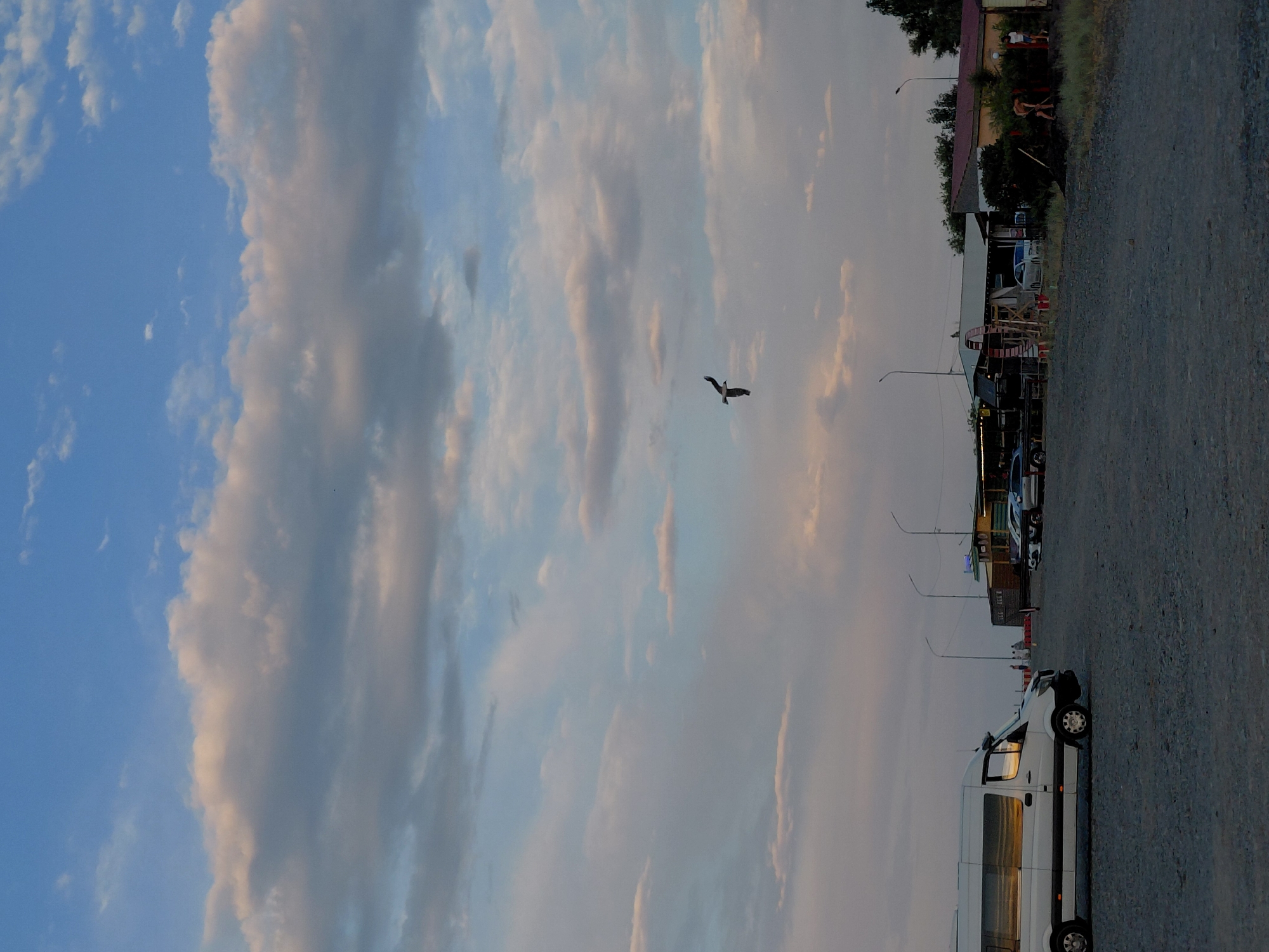 The image depicts a scene in Ақши, Kazakhstan, likely during the late afternoon or early evening. The sky is filled with a mix of blue and dramatic clouds, tinged with hues of pink and orange, suggesting a sunset or sunrise. A solitary bird is captured in flight against this sky.
Below the sky, the scene transitions to a ground-level view. A gravel parking area occupies the foreground. A white van is parked prominently on the right side of the frame, its reflective windows mirroring the sky. In the background, a row of buildings with awnings and various structures suggests a commercial or recreational area, possibly near a body of water, given the likely location. Several cars are parked in front of these buildings, and a few people are visible, some near the buildings and others further in the distance. Streetlights are also visible, hinting at the approaching evening. The overall atmosphere appears calm and serene, with the natural beauty of the sky contrasting with the man-made structures and vehicles. No visible text is discernable in the image.