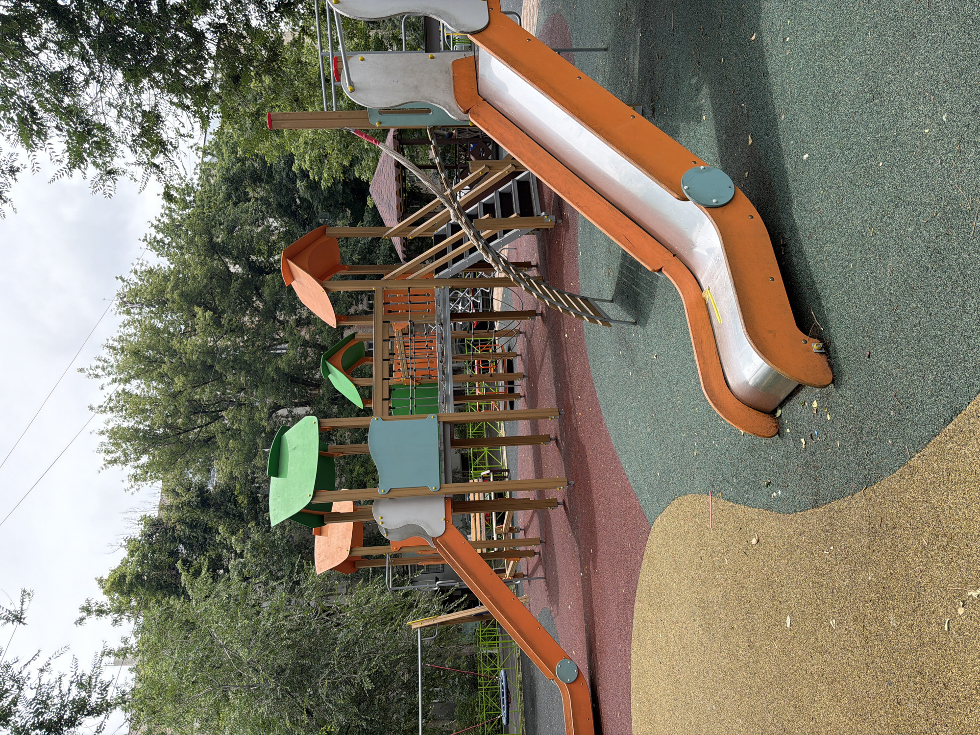 The image shows an outdoor playground with various play structures. The main focus is a large, multi-level wooden and orange plastic play structure with slides, climbing elements, and what appears to be a rope bridge. The ground is covered with a mix of green, red, and tan rubberized safety surfacing, with some loose leaves and debris scattered about. In the background, lush green trees create a natural backdrop, and the sky is overcast, suggesting a cloudy day.
The playground equipment includes a prominent orange slide with a silver chute, connected to a higher platform. Below this, there are additional levels with wooden beams, orange and green plastic panels, and a ladder. A rope bridge hangs between two sections of the structure. Further down, another orange slide is visible, curving towards the ground. The presence of a swing set frame can be seen in the lower left of the image, though no swings are attached.
The scene appears to be a public park or recreational area. The lighting suggests it is daytime, but the overcast sky indicates a lack of direct sunlight, possibly late morning or afternoon. There are no people visible in the image, and the playground equipment seems to be in good condition. The setting is clearly outdoors, amidst trees and under a cloudy sky. No visible text is present in the image. The location context is Shymkent, Kazakhstan.