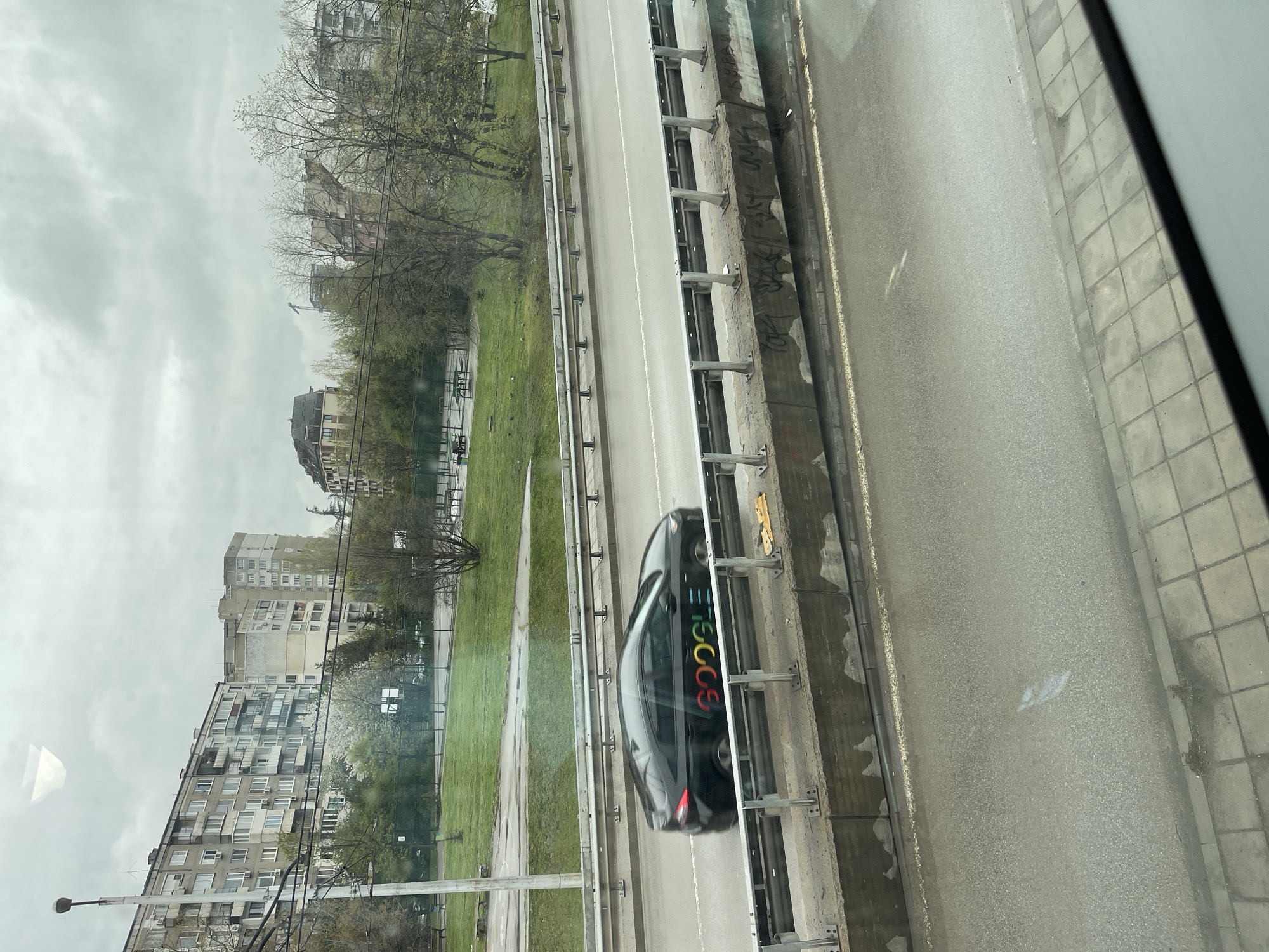 The image captures an urban landscape under an overcast sky, depicting a multi-lane road and surrounding environment. A black car, distinctively marked with the multi-colored "GOOGLE" logo on its side, is the central subject, appearing to drive along the road.
The road is made of asphalt with clear white lane markings. To the immediate right, a concrete barrier, bearing some faded graffiti, separates the main roadway from a paved area or sidewalk. On the left side of the road, a metal guardrail defines the edge of a wide, unkempt grassy area. This green space is dotted with deciduous trees, many of which are bare, suggesting the season is either late winter or early spring.
Beyond the grassy expanse, several multi-story residential buildings are visible, characterized by their blocky construction and numerous balconies, indicative of architecture common in Eastern European cities like Sofia, Bulgaria. In the distance, a construction crane can be seen. The sky is uniformly grey and cloudy, contributing to a muted, daytime lighting condition. No individuals are visible in the scene. The image provides a glimpse of a Google mapping vehicle operating within a residential urban setting.