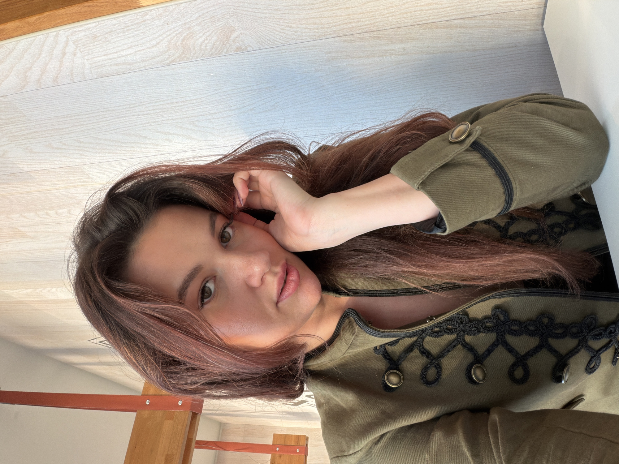 A young woman with long, dark brown hair highlighted with reddish/auburn tones is depicted in a close-up portrait. Her head is tilted to the side, and her right hand is gently placed against her cheek, with her fingers lightly touching her temple. She has fair skin, natural makeup, and her lips are slightly parted. Her dark eyes are looking towards the camera.
She is wearing an olive green jacket adorned with intricate black cord braiding and brass-colored buttons, giving it a distinctive military or vintage-inspired style. The jacket has details like a buttoned cuff on the left arm.
The background consists of light-colored wooden floorboards or paneling, with a section of reddish-brown wooden furniture, possibly a bed frame or shelving unit, visible in the lower left portion of the image. A white surface or wall is seen on the right. The image is oriented sideways, with the woman's head facing towards the left of the frame.
The lighting is soft and even, suggesting an indoor setting during the daytime. Her expression appears calm and natural. No visible text is present in the image. The scene takes place in Belgrade, Serbia.