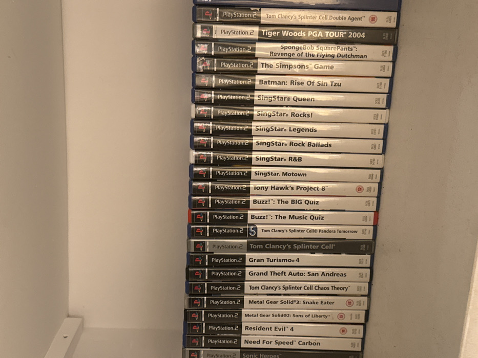 An indoor photograph captures a collection of approximately 24 PlayStation 2 (PS2) video game cases arranged vertically on a white shelf. The game cases predominantly feature black spines, each displaying the "PlayStation 2" logo at the top. Visible titles, from left to right, include 'Sonic Heroes', 'Need For Speed: Carbon', 'Resident Evil 4', 'Metal Gear Solid 2: Sons of Liberty', 'Metal Gear Solid 3: Snake Eater', 'Tom Clancy's Splinter Cell Chaos Theory', 'Grand Theft Auto: San Andreas', 'Gran Turismo 4', 'Tom Clancy's Splinter Cell', 'Tom Clancy's Splinter Cell: Pandora Tomorrow', 'Buzz!: The Music Quiz' (with a red spine), 'Buzz!: The BIG Quiz' (also with a red spine), 'Tony Hawk's Project 8', 'SingStar: Motown', 'SingStar: R&B', 'SingStar: Rock Ballads', 'SingStar: Legends', 'SingStar Rocks!', 'SingStar Queen', 'Batman: Rise Of Sin Tzu', 'The Simpsons Game', 'SpongeBob SquarePants: Revenge of the Flying Dutchman', 'Tiger Woods PGA TOUR 2004', and 'Tom Clancy's Splinter Cell Double Agent'. The games are uniformly aligned on the shelf, suggesting organized storage. The shelf surface is white, and the wall above it is also plain white, indicating a simple, clean interior environment. Artificial lighting illuminates the scene, casting soft shadows. No individuals or direct actions are depicted. The specific location within the city of London, United Kingdom, cannot be determined from the visual information provided in the image.