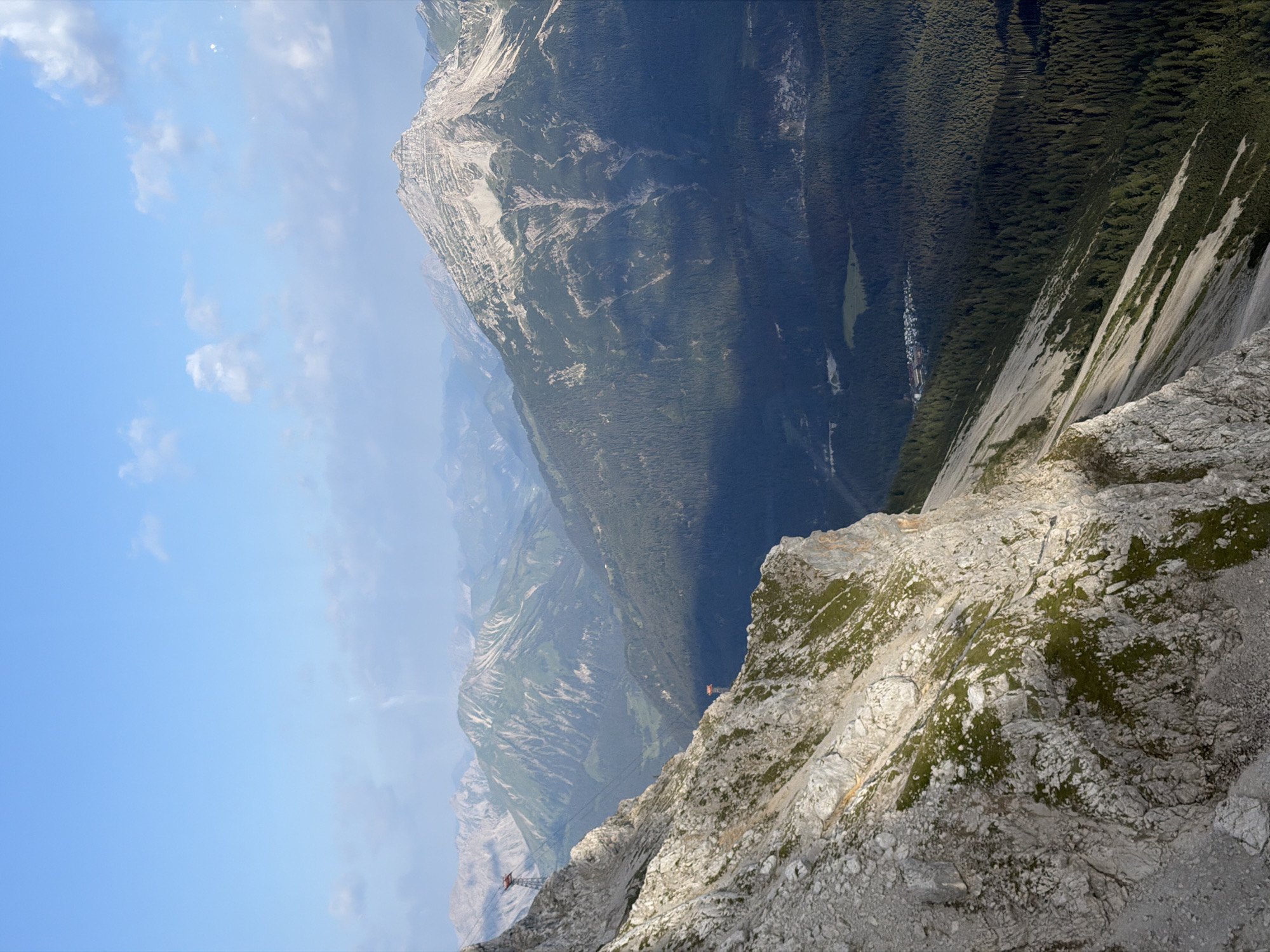 The image depicts a mountainous landscape with a clear blue sky dotted with scattered clouds. In the foreground, a steep, rocky cliff face with patches of green vegetation dominates the left side of the frame. To the right and in the background, a series of undulating mountains covered in dense dark green forests recede into the distance. The sun casts shadows across the slopes, highlighting the varied terrain. A few small structures, possibly buildings or a small village, are visible in the valley below, nestled among the trees. The overall scene conveys a sense of natural grandeur and remote wilderness. This location is identified as being in the vicinity of Grainau, Germany, a town known for its proximity to the Alps and its scenic beauty.
