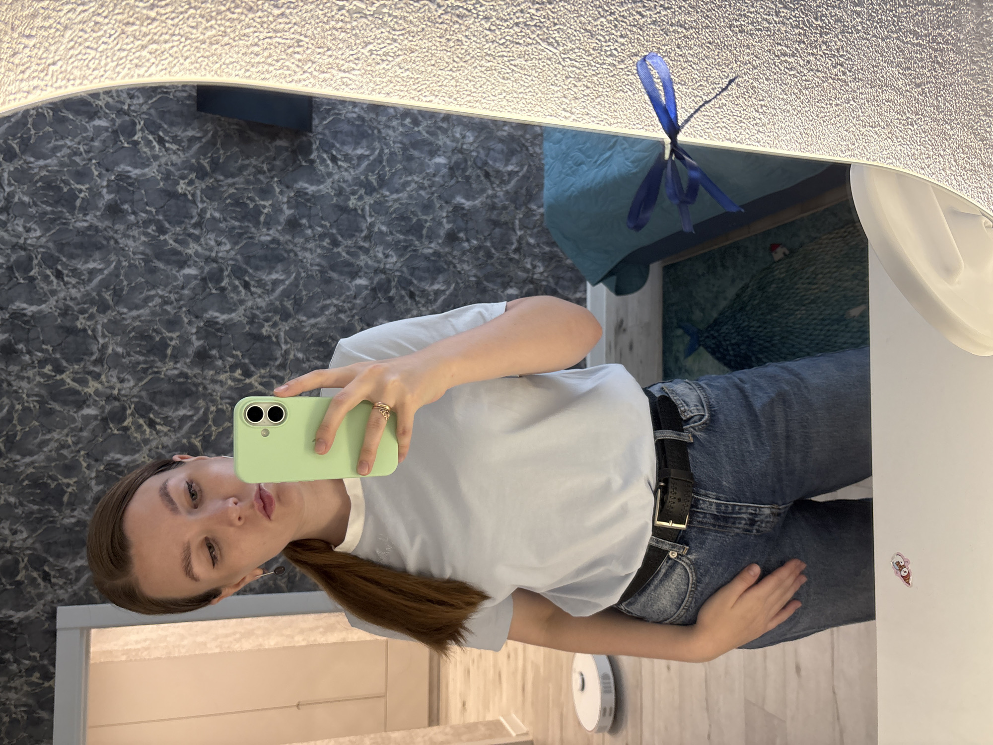 A young woman with brown hair tied in a ponytail is taking a selfie in a mirror. She is wearing a light blue t-shirt, blue jeans, and a black belt with a gold buckle. She is holding a mint green smartphone in her right hand. The background of the image shows a wall with a marble-like pattern, a doorway, and a robot vacuum cleaner on the floor. In the reflection of the mirror, a blue blanket and a piece of furniture are visible. The scene appears to be an indoor setting, likely a home, in the city of Izhevsk, Russia.
