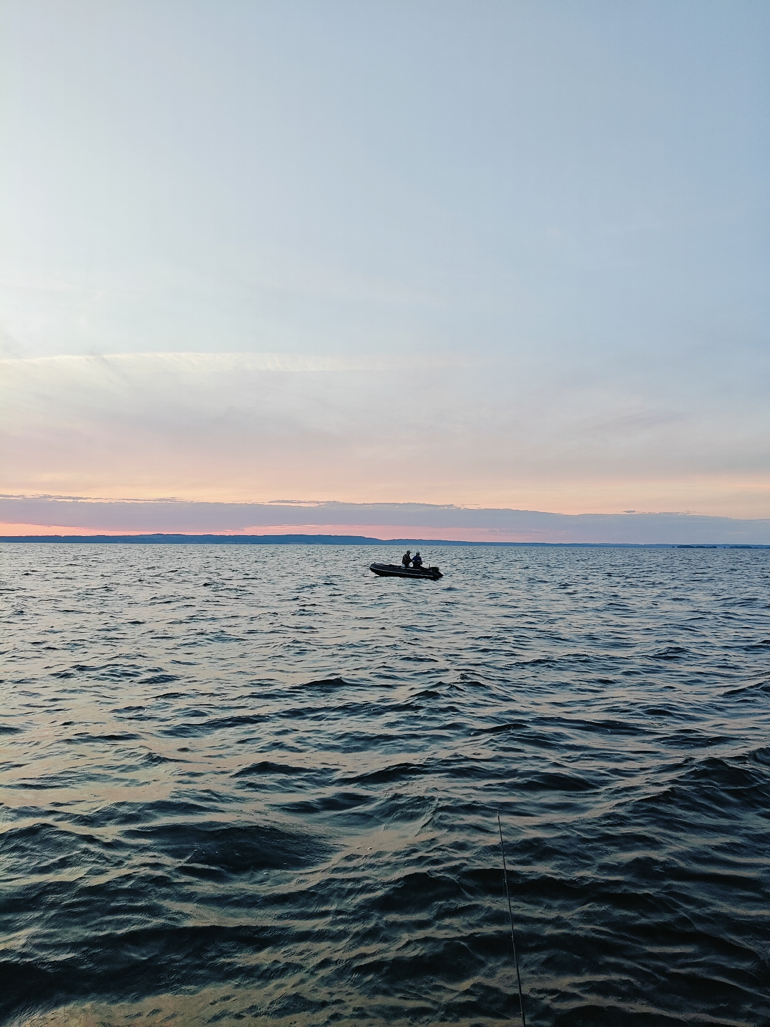 The image depicts a serene, open body of water, likely a lake or large river, under a pale twilight sky. A small, dark inflatable boat with two silhouetted figures is visible in the distance, centered horizontally and positioned slightly above the waterline. The water exhibits gentle ripples, reflecting the muted colors of the sky. The horizon line is low and indistinct, separating the water from a distant, hazy landmass. The foreground shows the texture of the water up close, with subtle reflections and dark hues. A fishing rod is visible in the lower right corner, angled upwards from the water's surface. The overall atmosphere is peaceful and calm, suggesting a late evening or early morning setting. The image has no discernible features that would allow for identification of the location within Stepanovo, Russia.