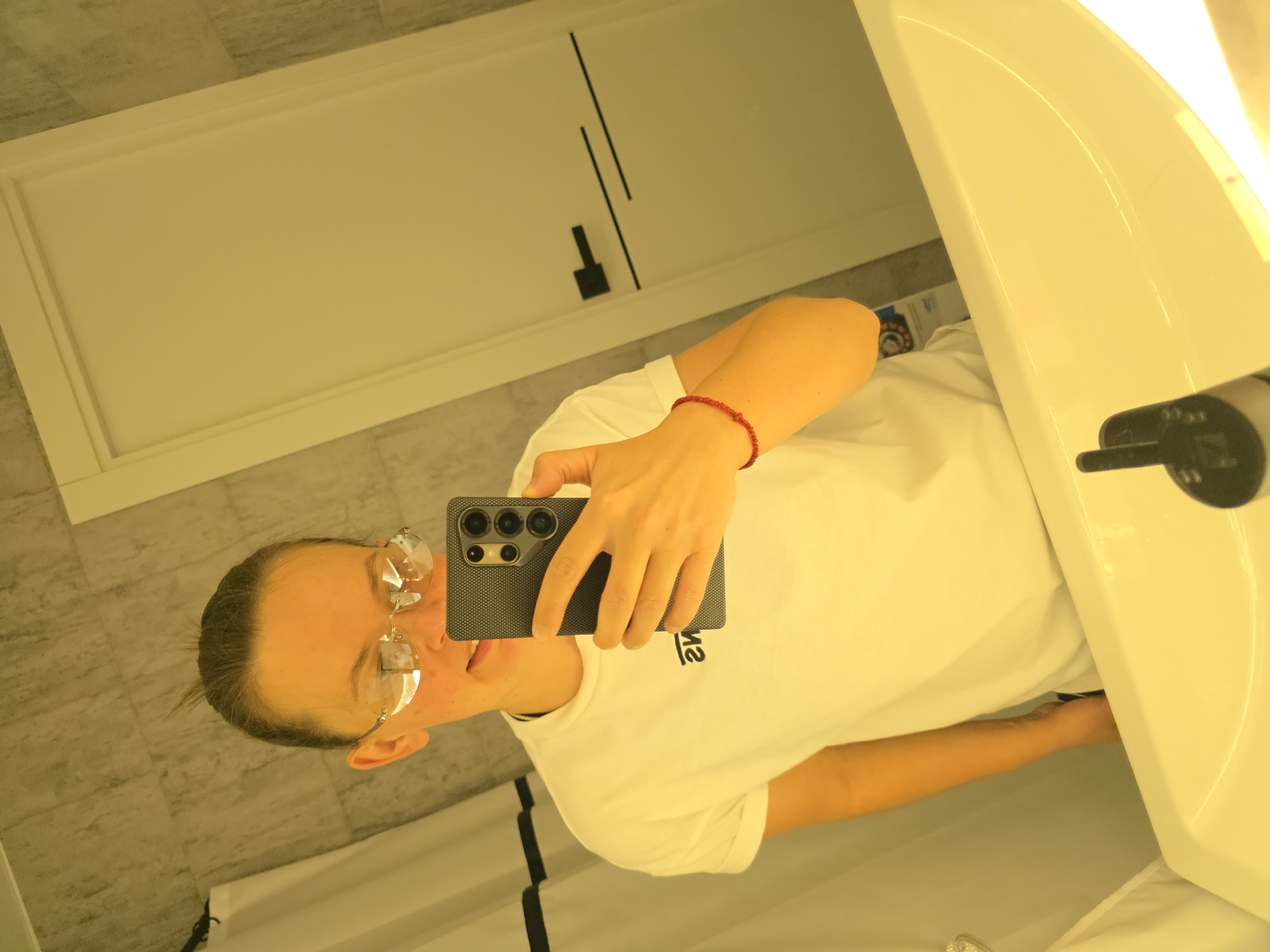 A woman stands in a bathroom, taking a selfie in a mirror. She is wearing a white t-shirt, glasses, and a red beaded bracelet on her right wrist. Her hair is pulled back in a bun. She is holding a black smartphone in her right hand. The bathroom has beige tiled walls and a white sink. A white shower curtain is visible to the left, and a white door with black accents is on the right. The image appears to have been taken in Rostov-on-Don, Russia.