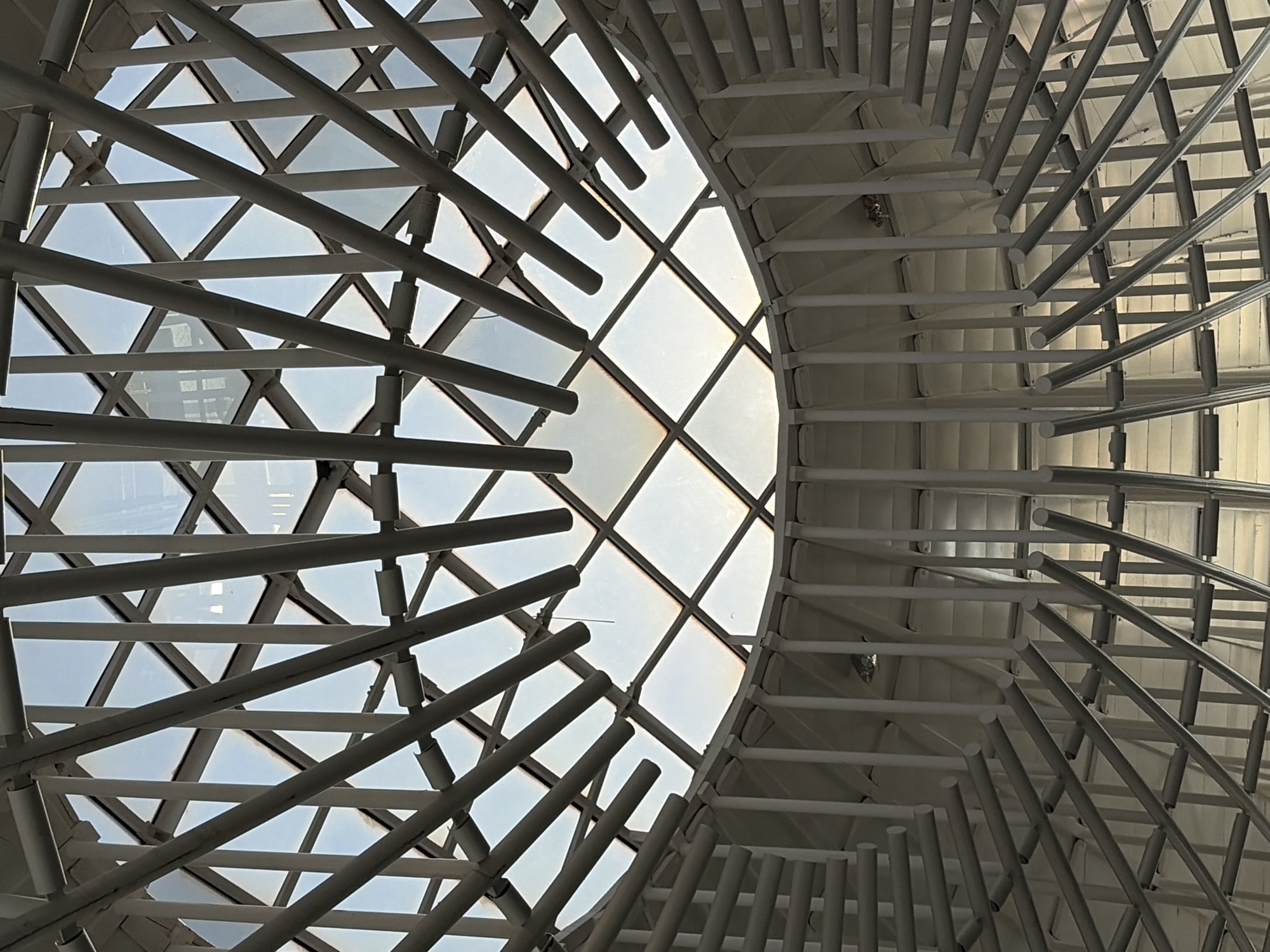 The image displays an intricate architectural ceiling or roof structure, viewed from below and bathed in natural light. The primary subjects are numerous light-colored, possibly white or silver, structural beams and support elements, along with glass panels forming a skylight.
On the left side of the frame, a dense grid of intersecting beams creates a complex geometric pattern, with clear sky visible through the triangular and square glass panels. Towards the center and right, the structure evolves into a more dynamic and curvilinear design. A large, approximately circular or oval opening is visible, also covered by glass panels, and surrounded by a dense arrangement of vertical and angled beams that appear to spiral or radiate outwards. These elements create a striking contrast between the angular framework on the left and the more fluid, organic forms on the right.
The bright light filtering through the skylight indicates it is daytime with a clear sky. No people or activities are visible, and there is no discernible text. The scene presents a modern, sculptural interior architecture, designed to maximize natural illumination. The building is located in Ciudad Guayana, Venezuela.
