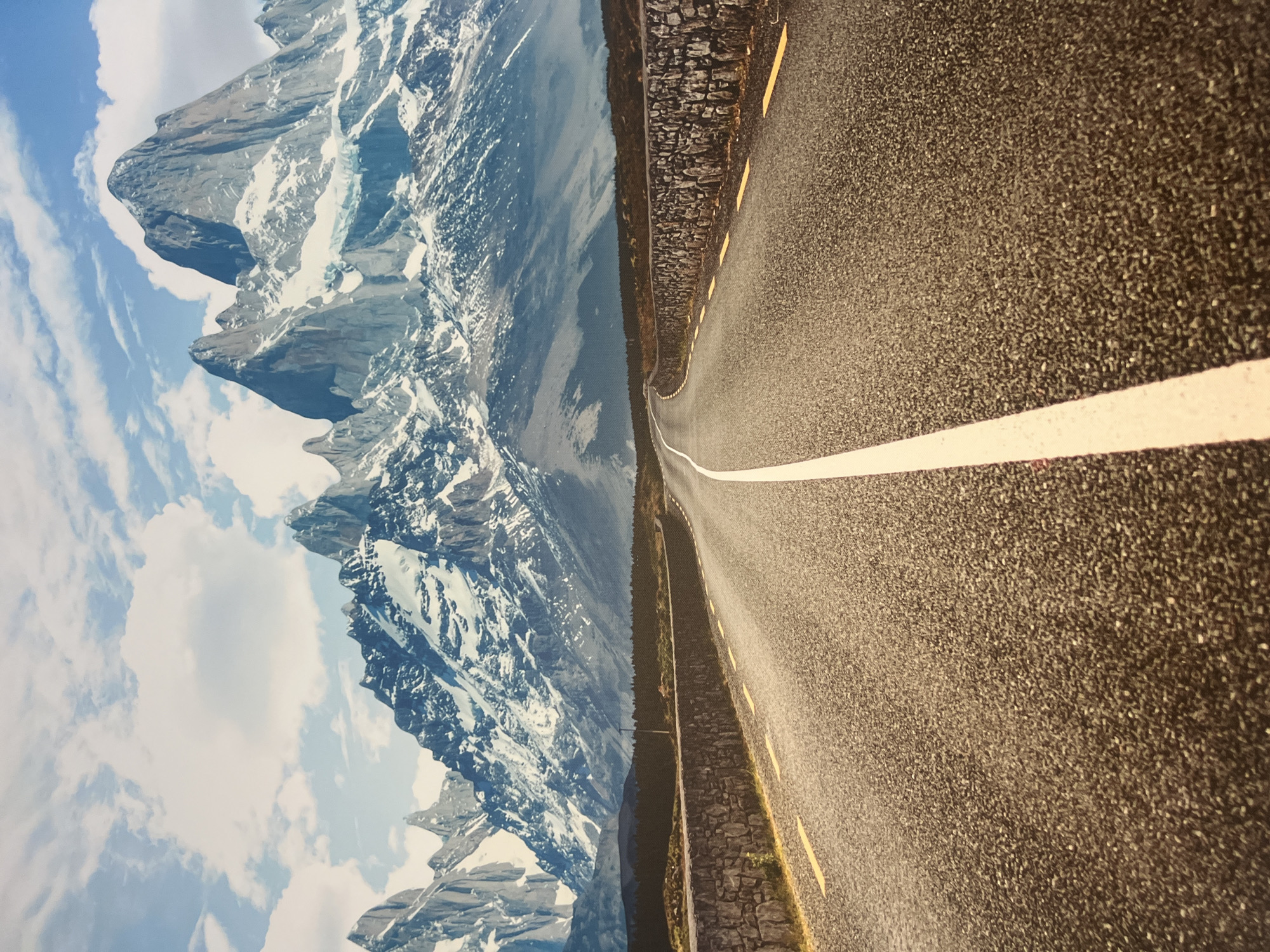 This media file depicts a winding asphalt road bordered by a stone wall on one side and majestic, snow-capped mountains on the other. The road features a white center line and intermittent yellow dashes along the edge. The mountains are rugged, with sheer rock faces and patches of snow, suggesting a high altitude. The sky is blue with scattered white clouds, indicating fair weather. The scene is oriented vertically, with the road and mountains appearing to ascend or descend. The lighting suggests it is daytime. There is no visible text in the image. The location is identified as Minzag, Russia.