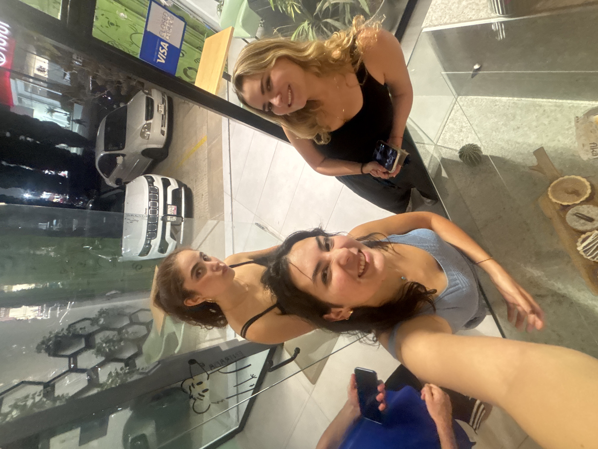 This media file presents a group selfie taken by three women inside a modern establishment with extensive glass windows, likely a cafe or shop, during nighttime. The image is angled to capture both the subjects and reflections of the exterior street scene.
Three women are featured, all displaying smiles. In the upper right, a blonde woman in a black tank top looks towards the camera while holding a smartphone. Below her, a woman with dark hair in a black-strapped top smiles subtly. The woman holding the camera and taking the selfie, in the foreground, has dark hair and wears a light blue ribbed top, smiling broadly directly at the lens. Another person, largely obscured and wearing a blue shirt, is partially visible below her, also holding a phone.
The interior setting features a light-colored tiled floor and a clean countertop. On the counter, a wooden board holds two small pastries or tarts, one unadorned and the other with white and chocolate stripes, next to a small potted cactus. A stylized drawing resembling Mickey Mouse is visible on the glass surface.
Reflected in the glass, the outdoor environment appears dark, illuminated by streetlights and building lights, confirming it is evening or night. Reflections show a street with parked vehicles, including a white SUV (possibly a BMW) and a white Jeep. Hexagonal planters are also visible through the window.
Notable visible text includes "VISA" and "CERRADO" (Spanish for "CLOSED") on a blue sign, a partial "Toyota" logo, "Jeep" on the vehicle's grill, and "aka" on a wrapper by the pastries. A partial "AUTO" is also seen in a reflection. The prevailing emotion among the subjects is happiness, conveyed through their smiles.