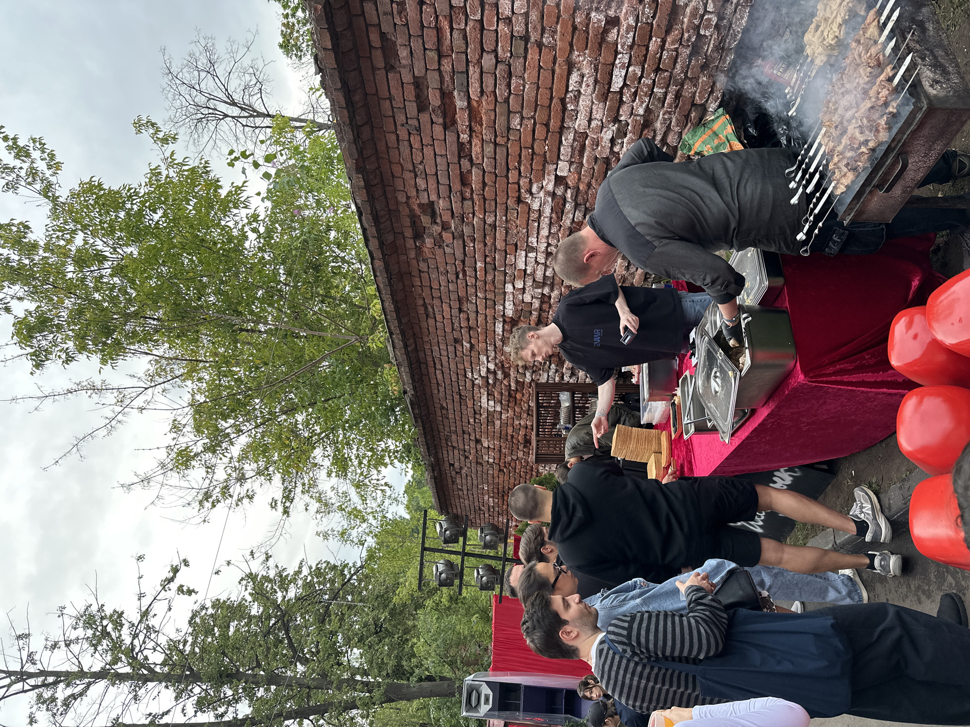 This media file captures an outdoor gathering, likely a barbecue or street food event, set against a backdrop of a weathered brick wall and lush green trees. Several people are visible, some actively engaged in preparing food and serving, while others form a queue or observe the proceedings.
The central focus is a large grill laden with skewers of meat, emitting visible smoke, indicating that food is actively being cooked. A table draped in a rich red velvet cloth displays various serving dishes and what appear to be stacks of disposable plates or containers. Two large, bright red, balloon-like objects are prominent on the right side of the frame, adding a pop of color to the scene. In the background, lighting equipment is set up, suggesting the event might extend into the evening or have a performance aspect.
The weather appears to be overcast, with a soft, diffused light. The presence of trees suggests an urban park or an outdoor venue. The casual attire of the individuals points to a relaxed, informal atmosphere. Emotions are not explicitly discernible, but the arrangement of people and the food preparation suggest a lively and communal event. No visible text is present in the image.