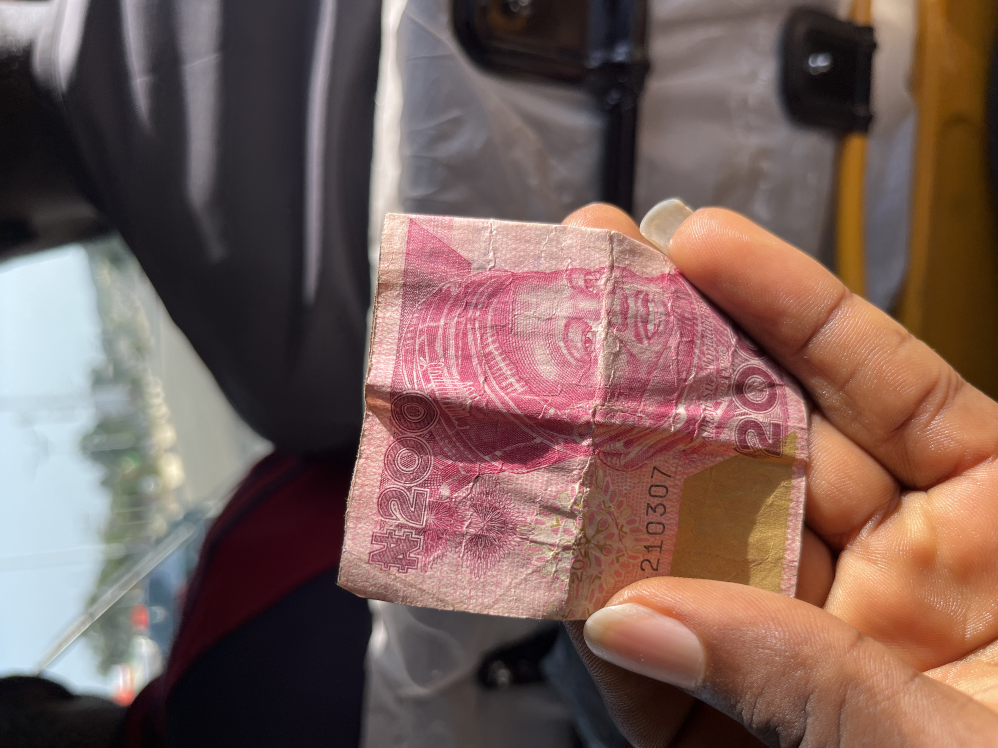 A close-up shot shows a person's hand holding a crumpled pink Nigerian 200 Naira banknote. The note has a serial number "210307" printed on it. The background is blurred but suggests an indoor setting, possibly the interior of a vehicle, with a white protective suit visible behind the hand and the currency. The lighting indicates it is daytime, with sunlight casting shadows. The scene appears to be in Bukuru, Nigeria. There are no explicit expressions of emotion visible.