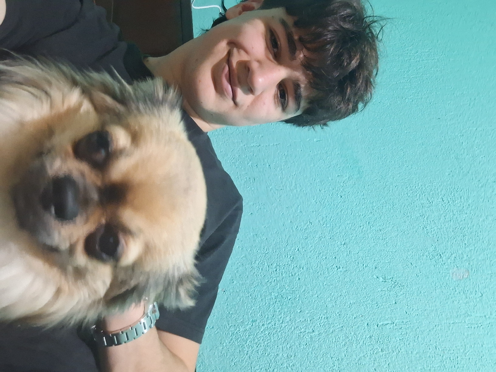 A young man with dark, curly hair and a light complexion smiles slightly while holding a small, tan and gray long-haired dog. The dog is positioned in the foreground, its face taking up a significant portion of the image. The man wears a dark-colored shirt and a silver watch on his wrist. A light blue wall serves as the background. The location in Stara Zagora, Bulgaria cannot be determined from this image.