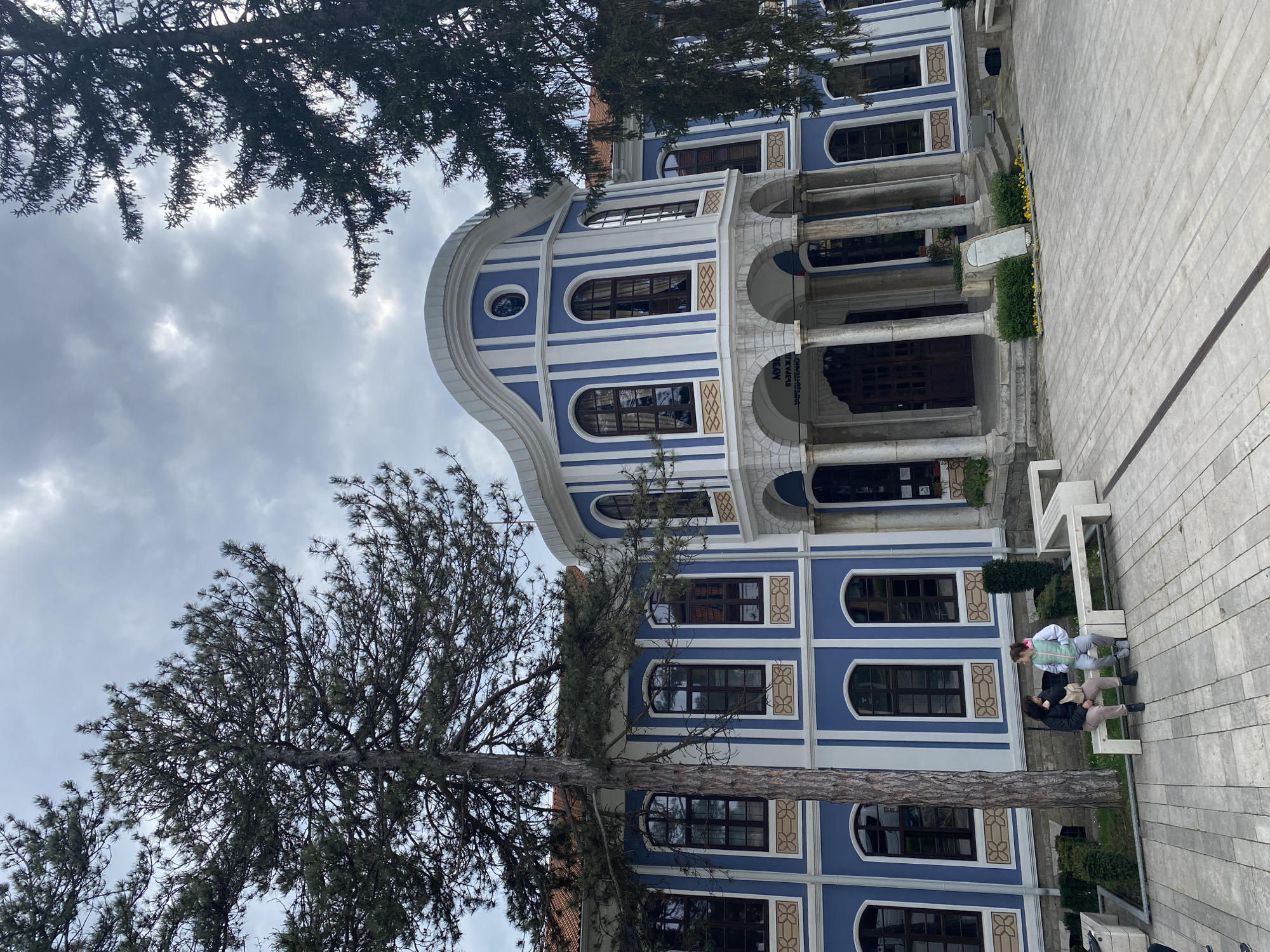 Here's a detailed description of the image:
**Location and Environment:**
The image shows the Regional Ethnographic Museum in Plovdiv, Bulgaria. The architectural style is distinctive, with a blue and white facade, arched windows, and a rounded upper section. The building stands on a paved area. Trees are visible framing the building on both sides. The sky is overcast with gray clouds.
**Subjects and Objects:**
The primary subject is the architectural structure of the museum. Additional details include the windows, columns, and ornate design elements of the building. Two people are sitting on a bench, situated in front of the museum. A tree grows in front of the building.
**People and Interaction:**
Two individuals are sitting on a modern, white bench in front of the museum. Their postures suggest they are engaged in conversation or simply resting.
**Action:**
The primary action is the presence of two individuals sitting and interacting on the bench. The building serves as the static backdrop to their interaction.