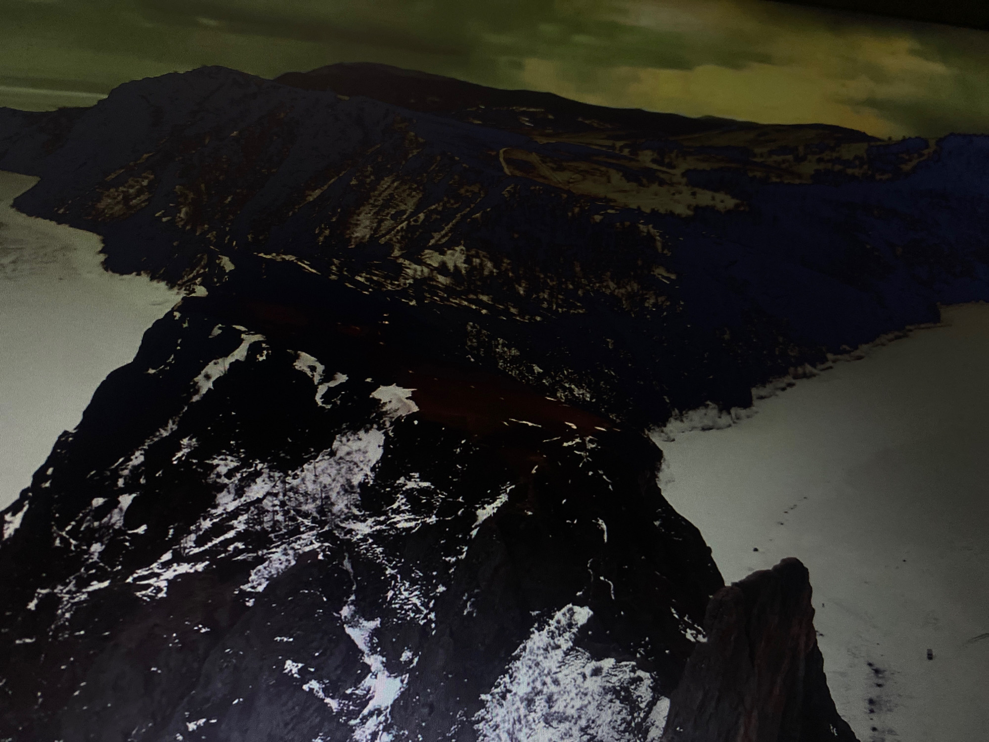 The image depicts a dramatic, high-angle aerial view of a rugged mountain range covered in snow and ice. The mountains are dark, with stark white snow clinging to their peaks and crevices. In the foreground, a prominent, jagged peak dominates the left side of the frame, casting a shadow across a vast, snow-covered plain that stretches into the distance. The plain appears to be a frozen body of water, possibly a lake or sea, with faint footprints or tracks visible, suggesting the presence of small figures or vehicles. Further back, more mountain ranges recede into the horizon, their slopes partially obscured by shadows and a muted, greenish-yellow sky. The overall impression is one of a remote, desolate, and frozen landscape. There is no discernible urban environment or any elements that would suggest the location of Hemayetpur, Bangladesh.