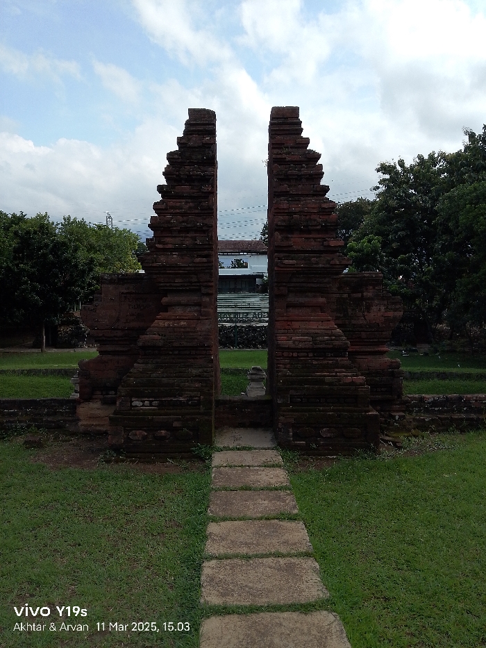 The image shows a stone archway made of brick, located at the coordinates -6.736, 108.543 in the city of Cirebon, Indonesia. The archway is made of layered bricks and is weathered with age. It is surrounded by green grass and leads to a walkway made of flat stone slabs. Trees line the outer edges of the archway. The sky is overcast with a hint of blue, indicating a partly cloudy day. The scene is serene and tranquil. There is a small structure in the center of the archway, which appears to be a simple, weathered monument. It is difficult to see the specifics of the monument, as its details are obscured by the distance and the low-resolution quality of the image. No people are present in the scene.