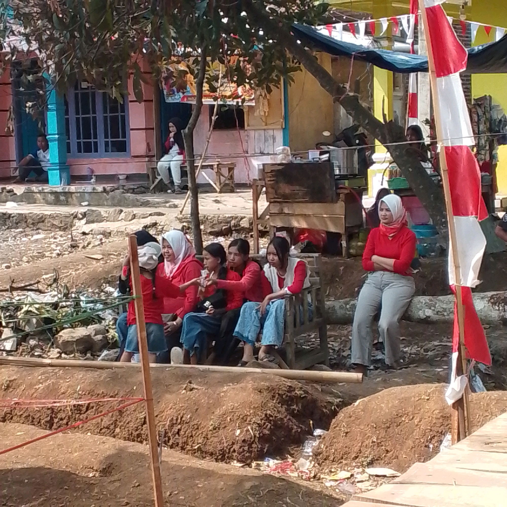In Cilaku, Indonesia, a group of people are gathered outdoors. In the foreground, there is a mound of disturbed earth, possibly a construction site or an excavation, with red string marking boundaries. A wooden post stands vertically near the edge of this disturbed area. Behind the mound, a group of young people, predominantly females, are seated on a wooden bench. Some are wearing red tops and blue jeans, while others have headscarves. One individual on the far right of this seated group, a woman wearing a white headscarf and a red long-sleeved shirt, is facing the viewer with her arms crossed. To the left of the bench, a person wearing a black headscarf and a red top is seated, holding what appears to be a small object. Further to the left, two individuals, a male and a female, are seated on a raised ledge in front of a building with a pink facade and blue-trimmed windows. The building has posters or signs on its exterior. In the background, more structures are visible, including a yellow building adorned with red and white triangular flags. A red and white flag on a pole is prominently displayed on the right side of the image. The environment appears to be a public space with elements of casual gathering and possibly ongoing activity.