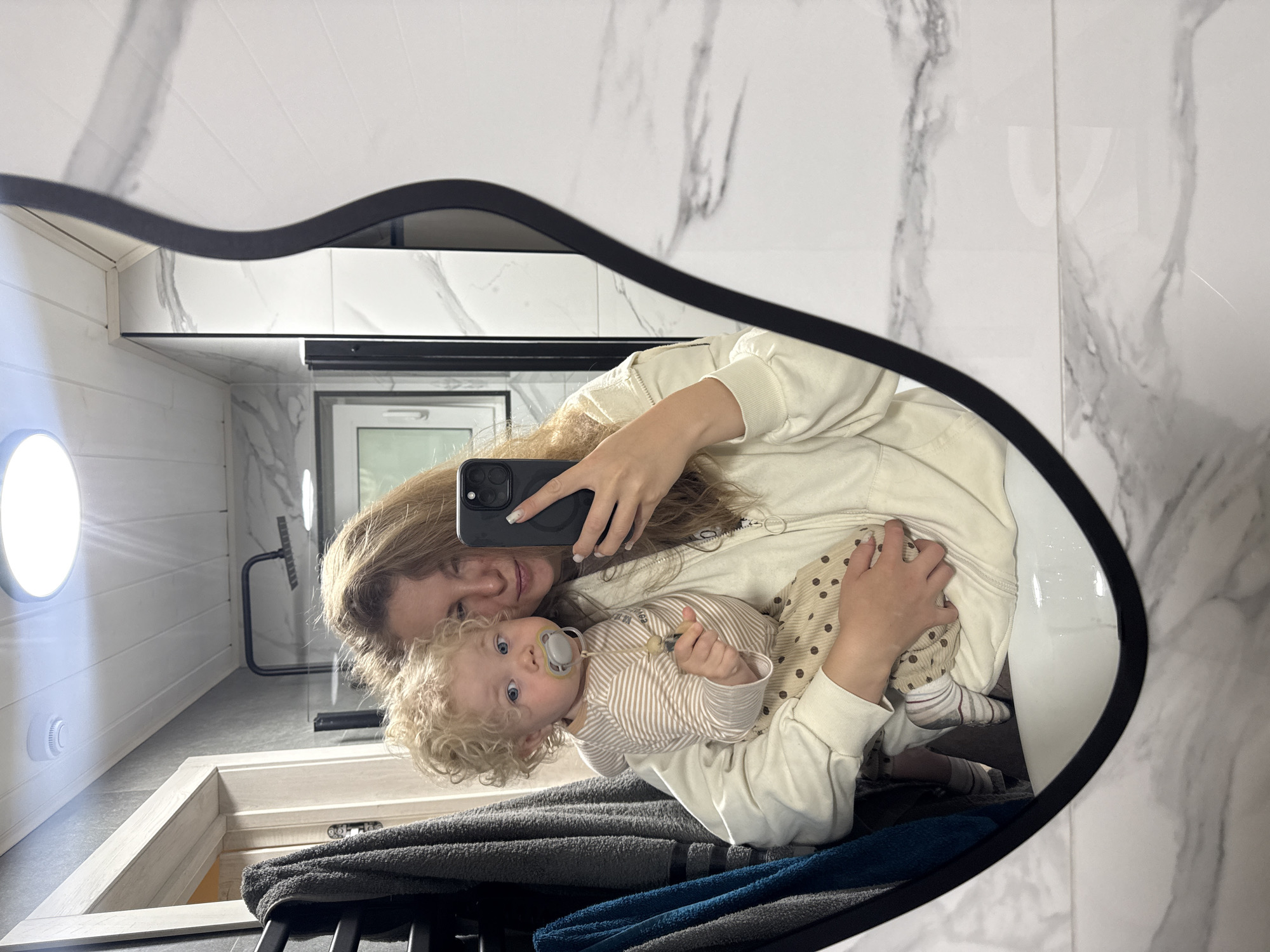 In a modern bathroom with white marble-patterned tiles, a woman stands in front of a mirror, taking a selfie. She is holding a baby with curly blond hair, who is wearing a pacifier and a striped long-sleeved shirt with polka-dot pants. The woman has her hair down and is wearing a cream-colored zip-up hoodie. A black iPhone is visible in her hand, held up to take the photo. In the reflection, a black towel rack with gray and blue towels is to the left, and a white sink is below. The bathroom ceiling has a circular light fixture. The image is framed by the wavy edge of the mirror. The location is Prokudino, Russia.