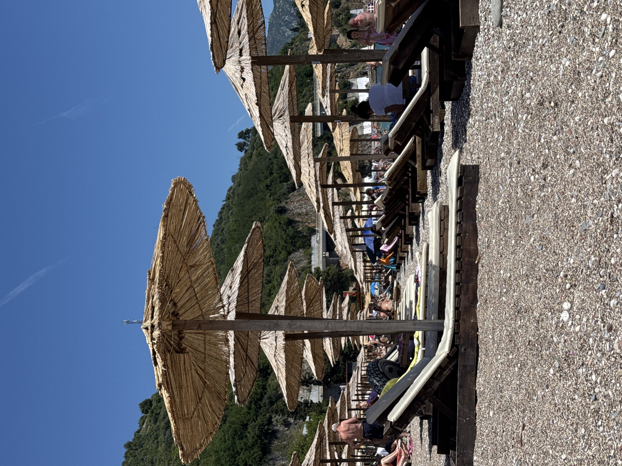 The image depicts a sunny day on a crowded pebble beach in Budva, Montenegro, likely located in the vicinity of Slovenska Plaža, a popular tourist area. The foreground is dominated by rows of empty and occupied sun loungers, many with thatched umbrellas providing shade. The beach is covered in a dense layer of small, multi-colored pebbles. In the midground, several people are visible relaxing on loungers or walking. Some are partially obscured by the umbrellas and loungers. In the background, lush green hills rise steeply, and a modern highway overpass is visible traversing the landscape. The sky is a clear, bright blue with a faint contrail from an aircraft. The overall impression is a typical busy beach scene on a warm summer day, with a blend of natural landscape and man-made infrastructure.