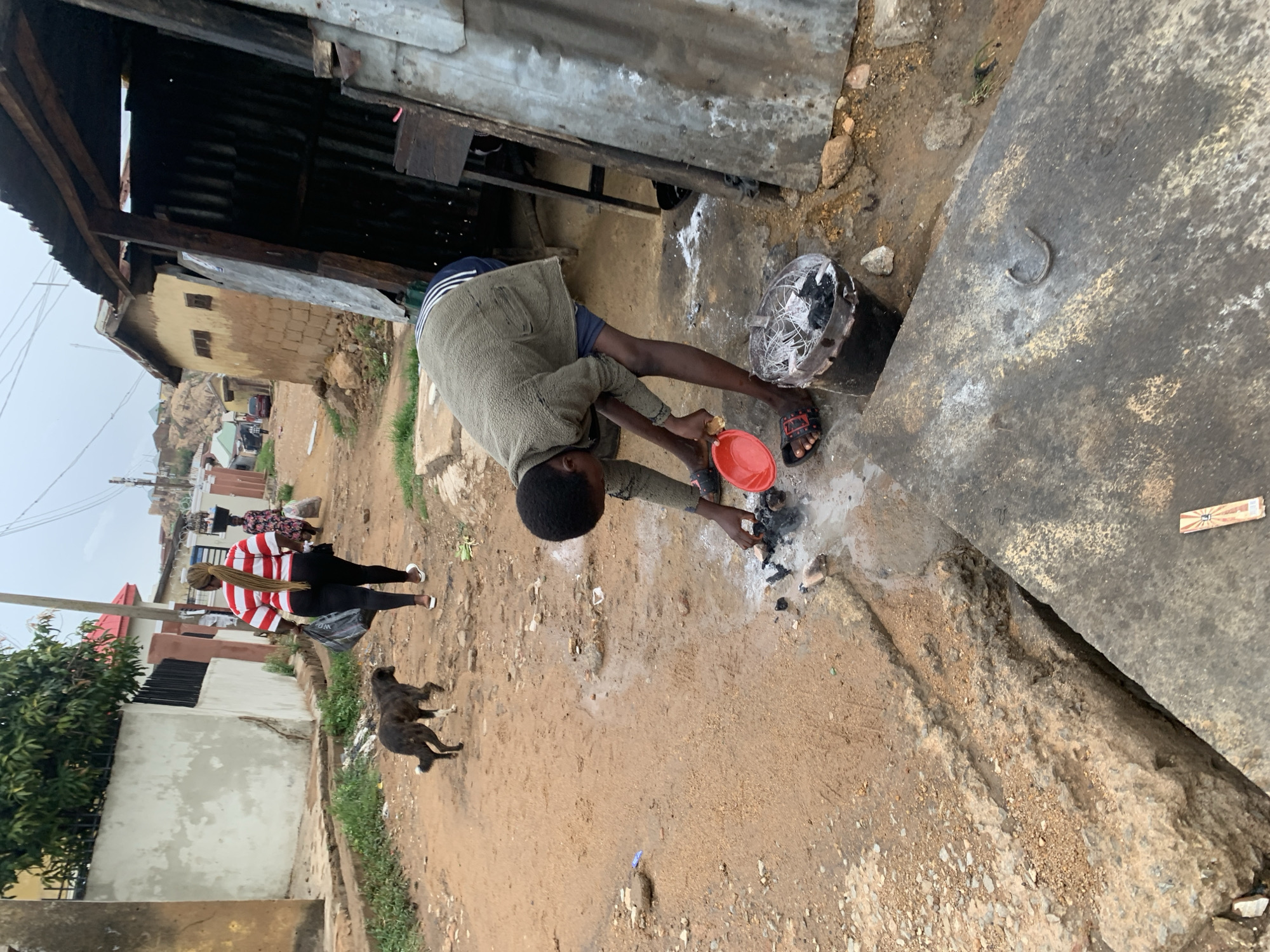 A young man is bending over a small fire on a dirt road in Jos, Nigeria, holding a red bowl. The man wears a green jacket, blue shorts, and red sandals. He appears to be tending to the fire with his right hand. Near the fire is a rusted, damaged fan. In the background, a woman wearing a striped shirt and black pants walks down the road, accompanied by a black dog. Various buildings and structures are visible in the background, including a tin-roofed shack, a yellow brick building, and a house with a red roof. The ground is uneven and appears to be made of dirt and gravel. A large, gray, concrete slab is positioned on the right side of the frame.