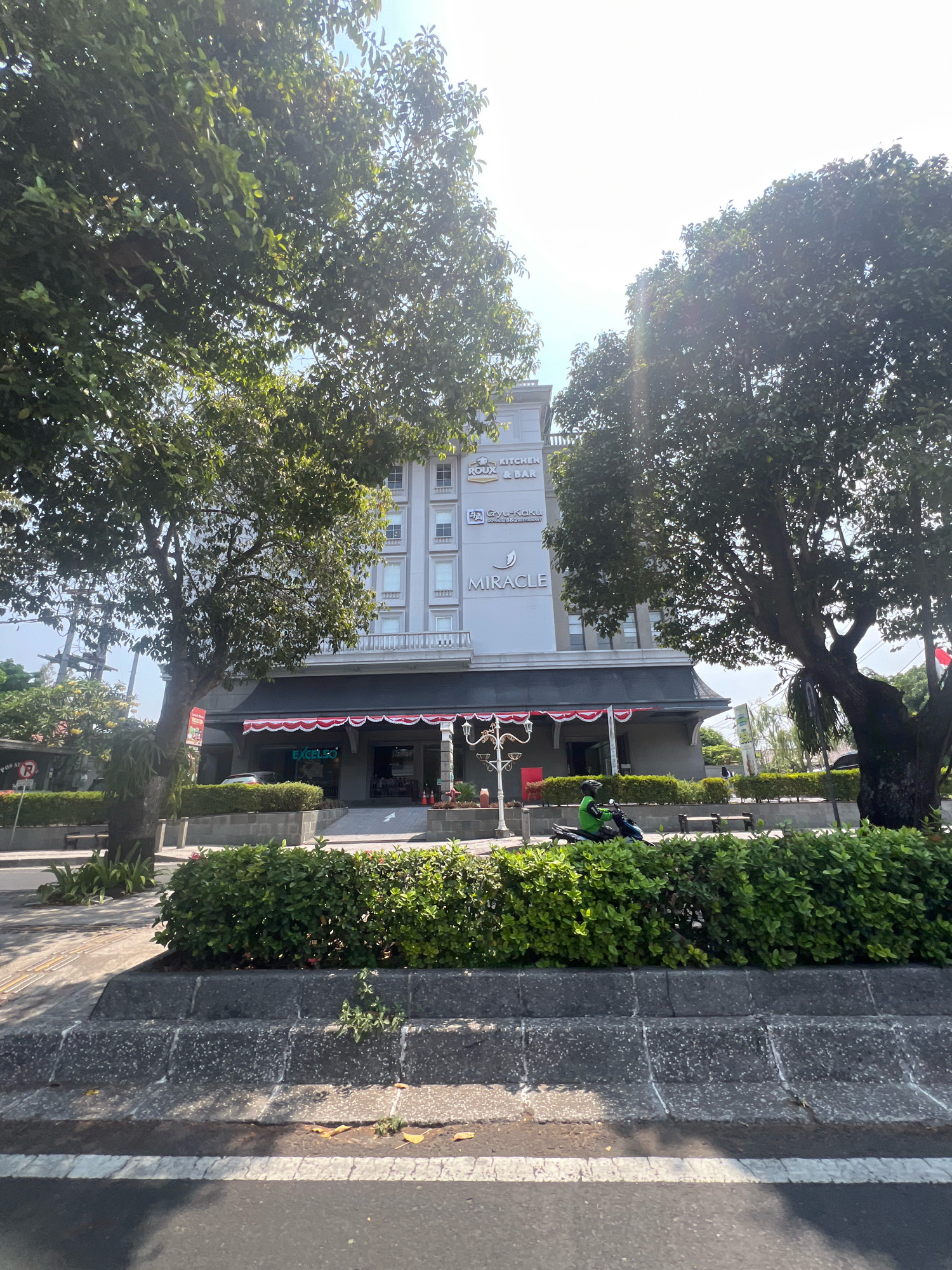 The image captures a street scene in front of the Miracle Building, located in Yogyakarta, Indonesia. The building, a multi-story structure with a modern facade, features prominent signage for "MIRACLE," "ROUX & BAR," and "GUY-KAKU." A portion of the ground floor is visible, with awnings and an entrance leading into a darkened interior. In front of the building, a neatly trimmed hedge forms a barrier between the sidewalk and the street. A person on a green motorcycle, wearing a helmet and possibly a uniform, is riding on the street, partially obscured by foliage. The surrounding environment includes trees, streetlights, benches, and a sign indicating a pedestrian crossing or traffic regulation. The lighting suggests it is daytime, with sunlight casting shadows. The overall scene depicts a typical urban environment with commercial activity.