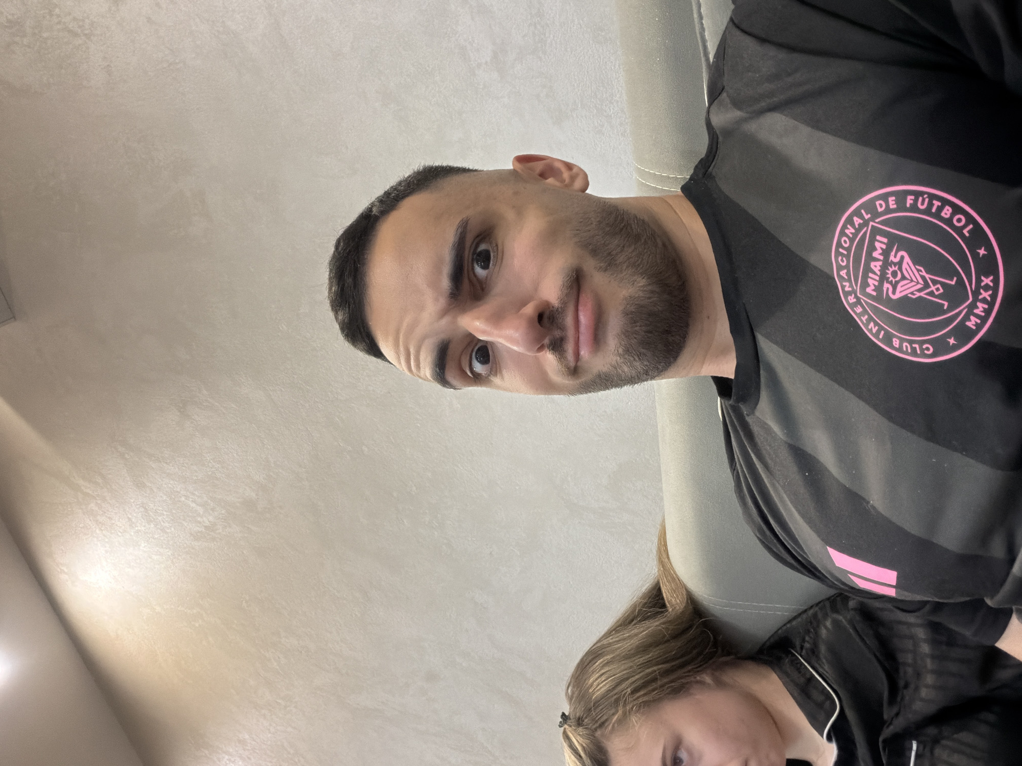 A close-up shot shows a man and a woman in what appears to be an indoor setting. The man, with a dark beard and short hair, is wearing a black Inter Miami CF soccer jersey with pink accents, indicating he is a fan of the team. The woman, with blonde hair, is partially visible and is wearing a black satin top. The background includes a textured wall and part of a grey sofa. There is no information indicating the location is Plovdiv, Bulgaria.